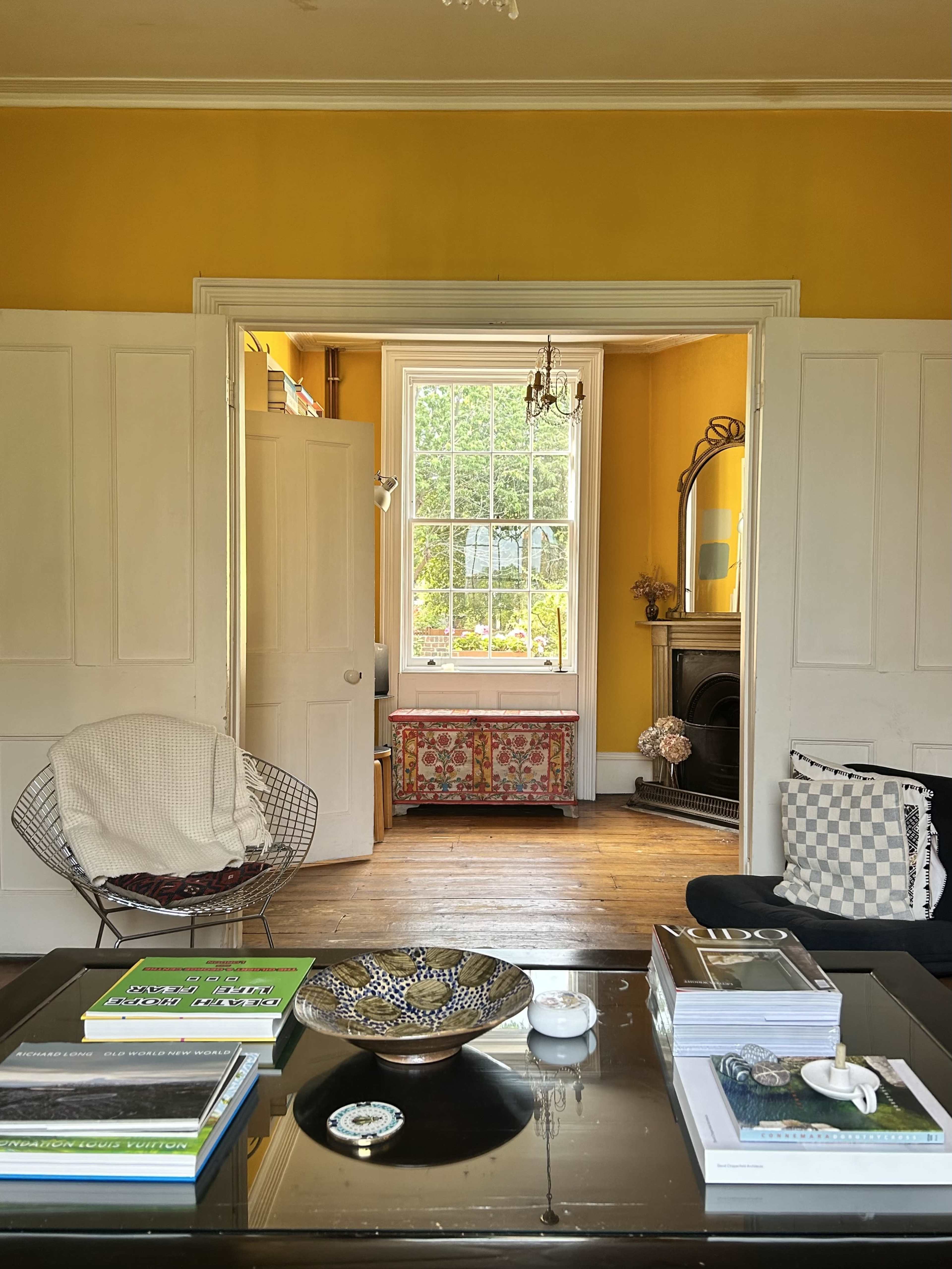 A living room features a black coffee table with books and decorative items, leading to a brightly lit hallway with yellow walls and a window overlooking a garden.