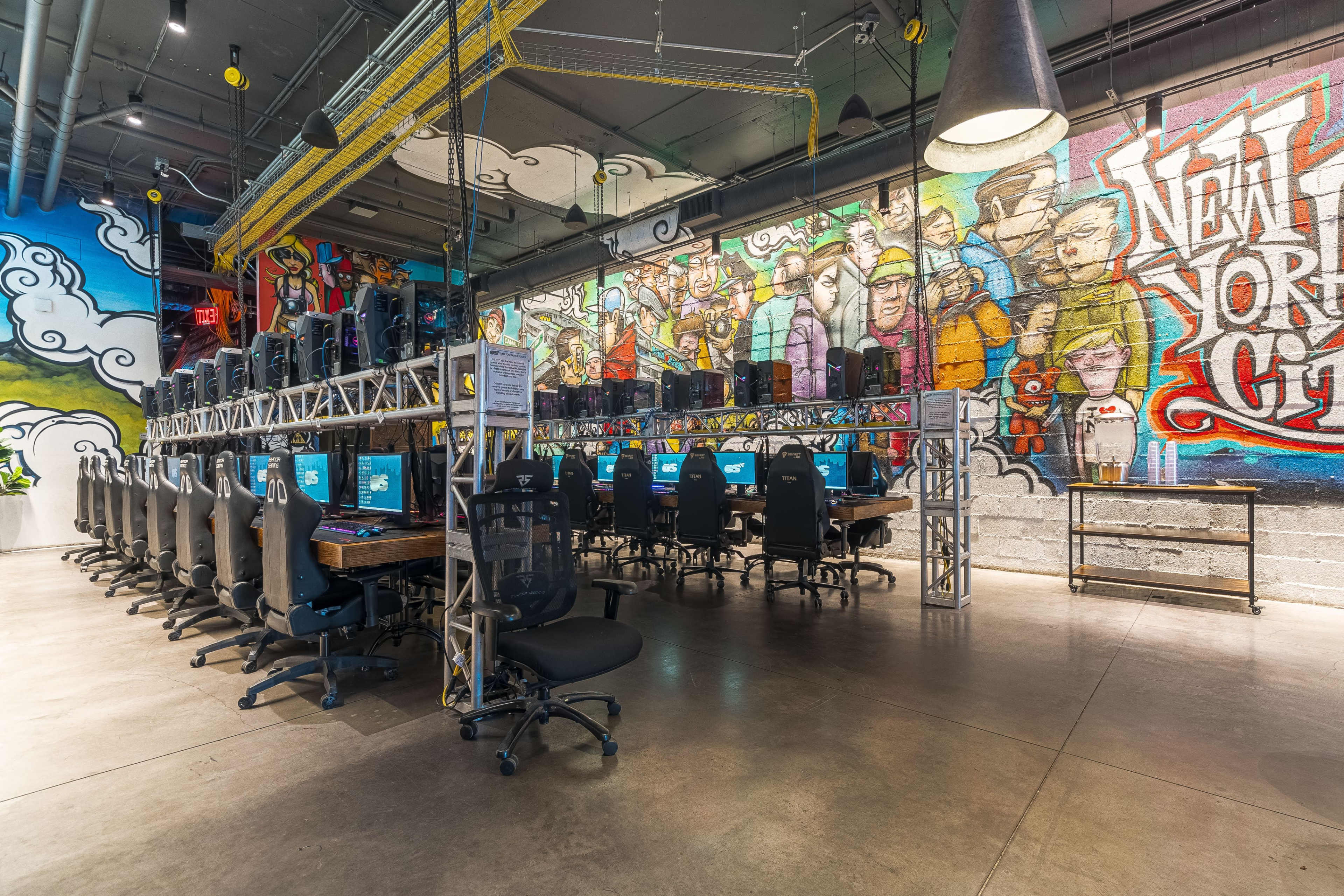 A modern coworking space featuring rows of computers arranged on metal desks beneath a colorful mural depicting urban life.