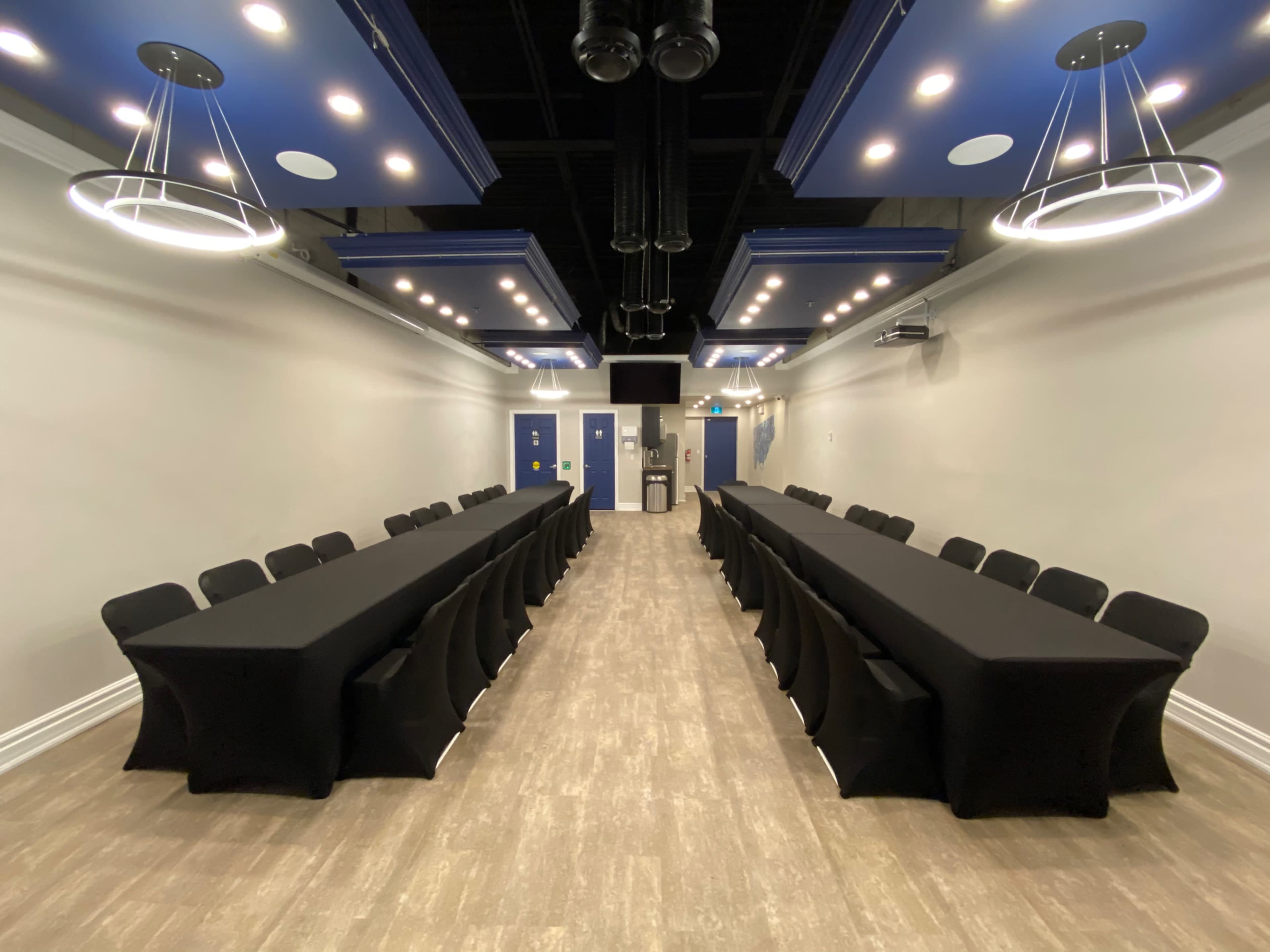 A conference room with two long black tables arranged in a U-shape, surrounded by black chairs, and illuminated by modern overhead lights.
