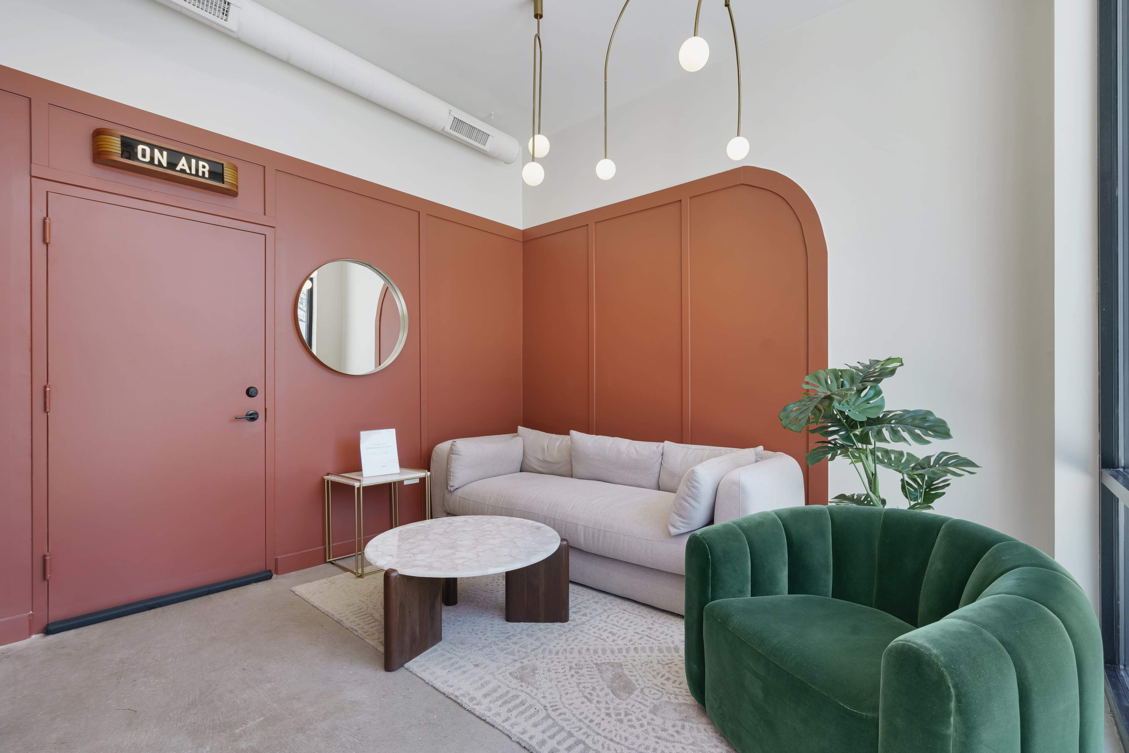 A modern lounge area featuring a pink and white color scheme, with a circular mirror, a green velvet chair, a light-colored sofa, a round coffee table, and a wall sign that reads "ON AIR."