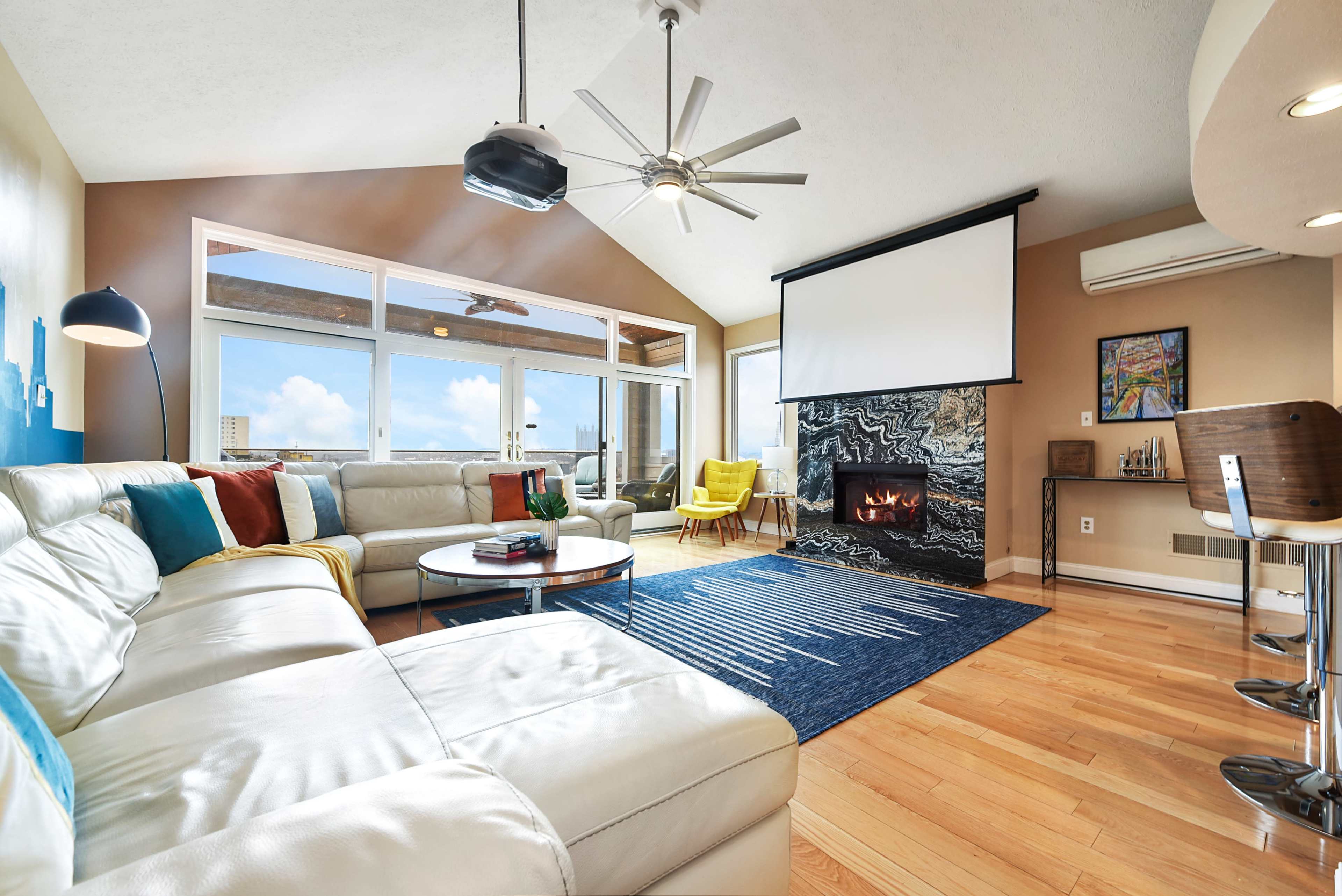 A modern living room features a sectional couch, a sleek coffee table, a large area rug, and a fireplace against a backdrop of large windows overlooking a cityscape.
