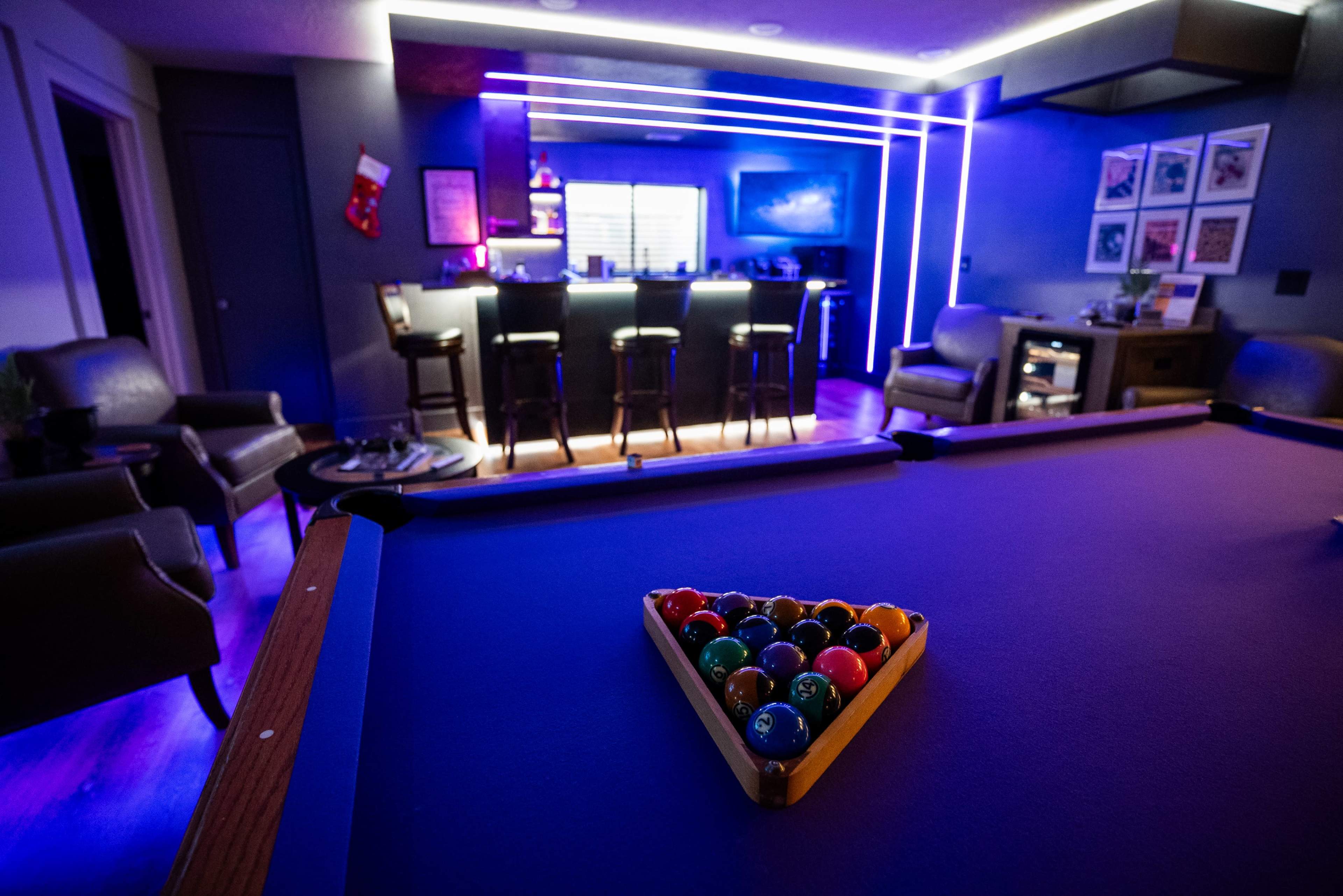 A triangular rack of billiard balls sits on a purple pool table in a dimly lit room with a modern bar and colorful lighting.