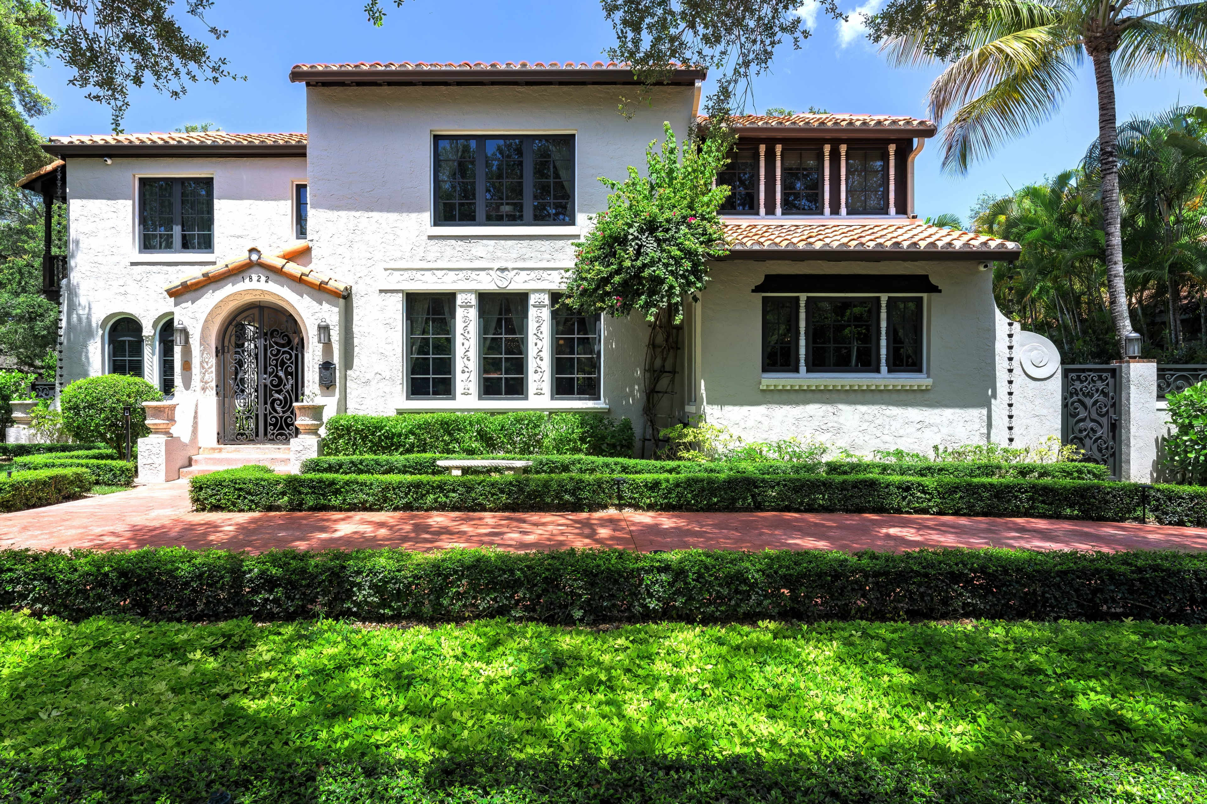 The image shows a two-story house with a white stucco exterior, terracotta roof, and manicured hedges, situated on a lush green lawn.