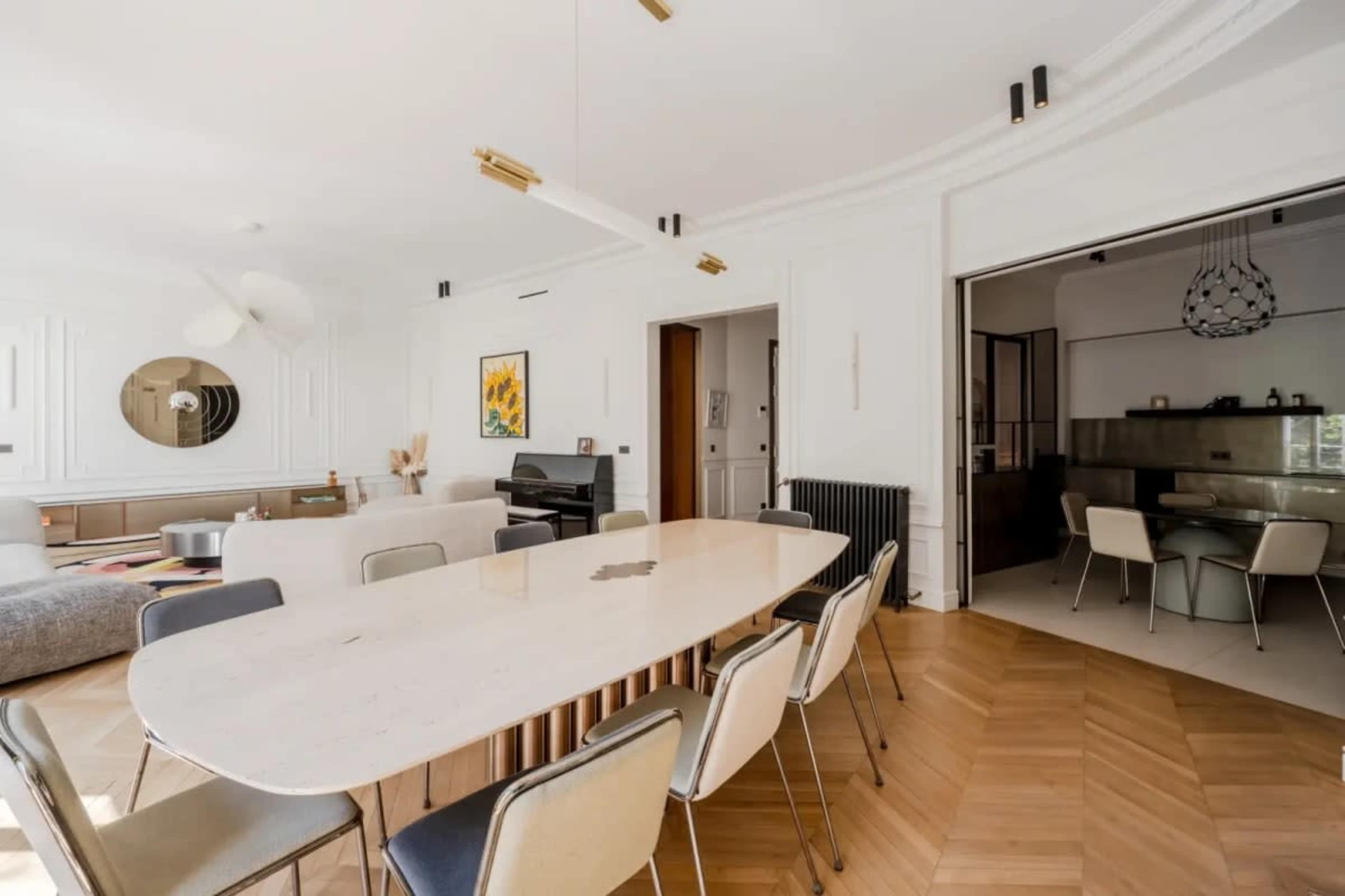 The image shows a modern dining area featuring a large, oval table surrounded by chairs, with a white wall and a visible open kitchen in the background.