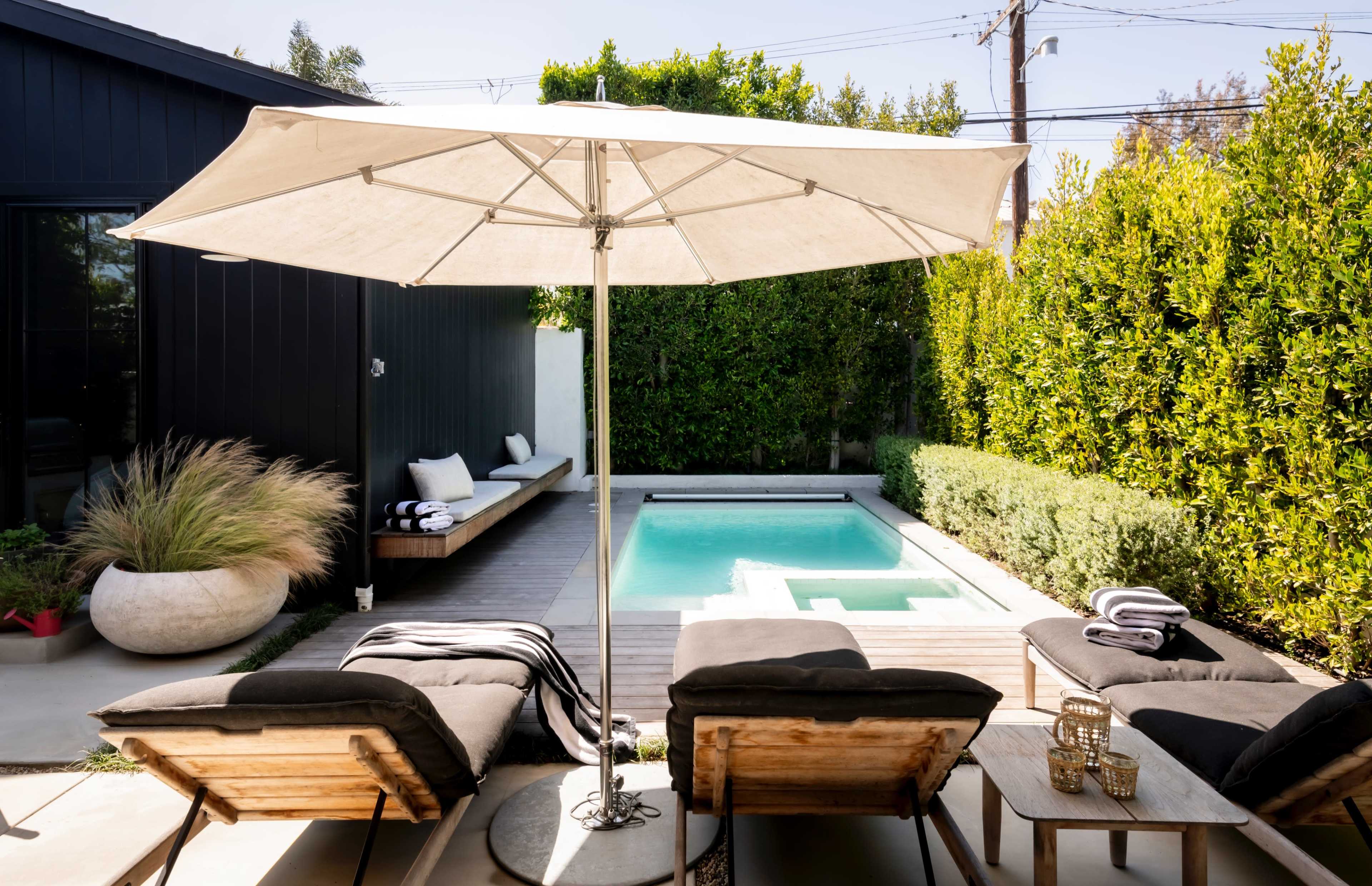 A poolside area features two lounge chairs, a large umbrella, and a small swimming pool surrounded by neatly trimmed hedges and a wooden deck.