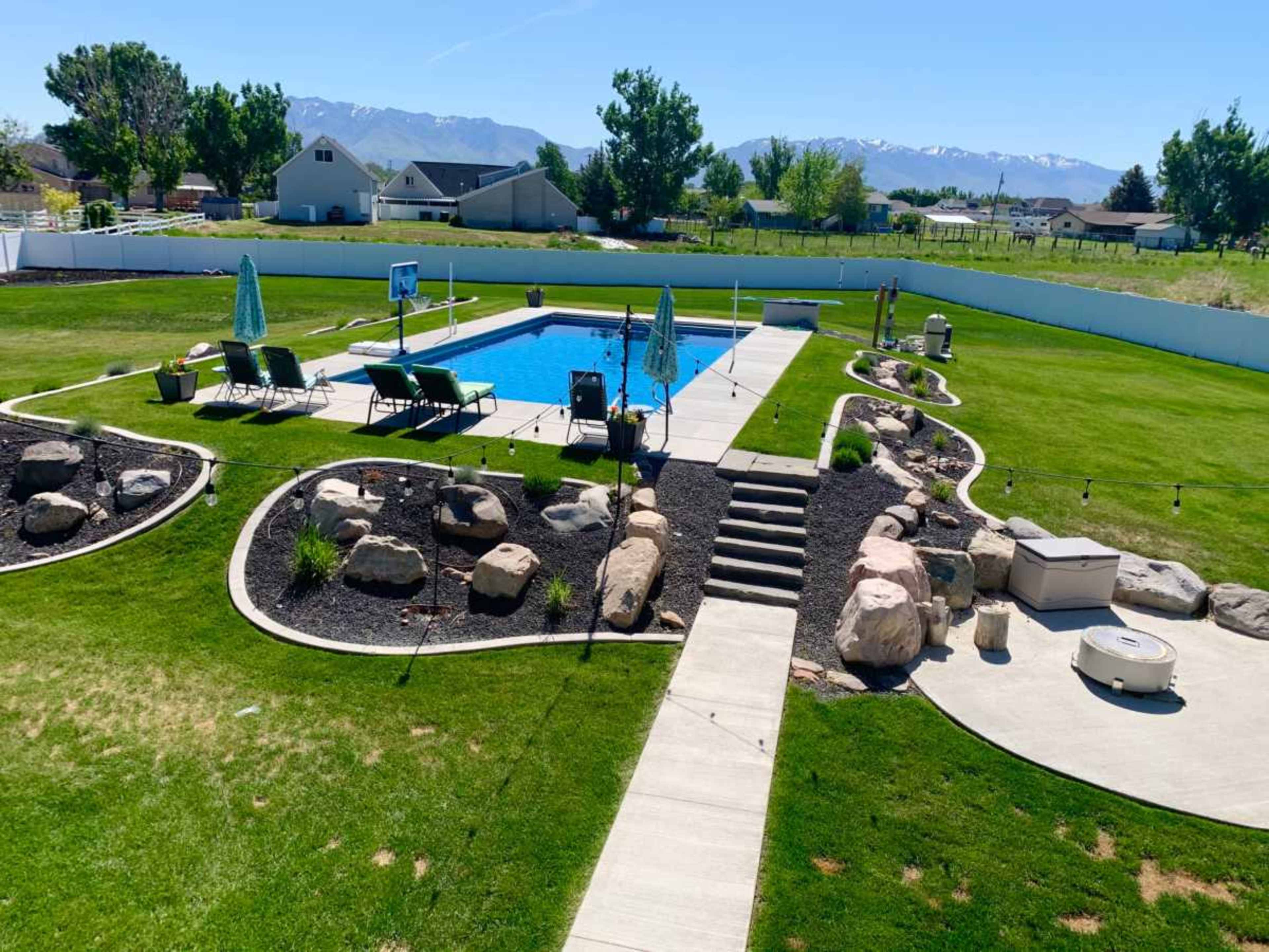The image shows a backyard pool area with a swimming pool surrounded by lounge chairs, landscaped rocks, and a neatly maintained lawn under a clear blue sky.