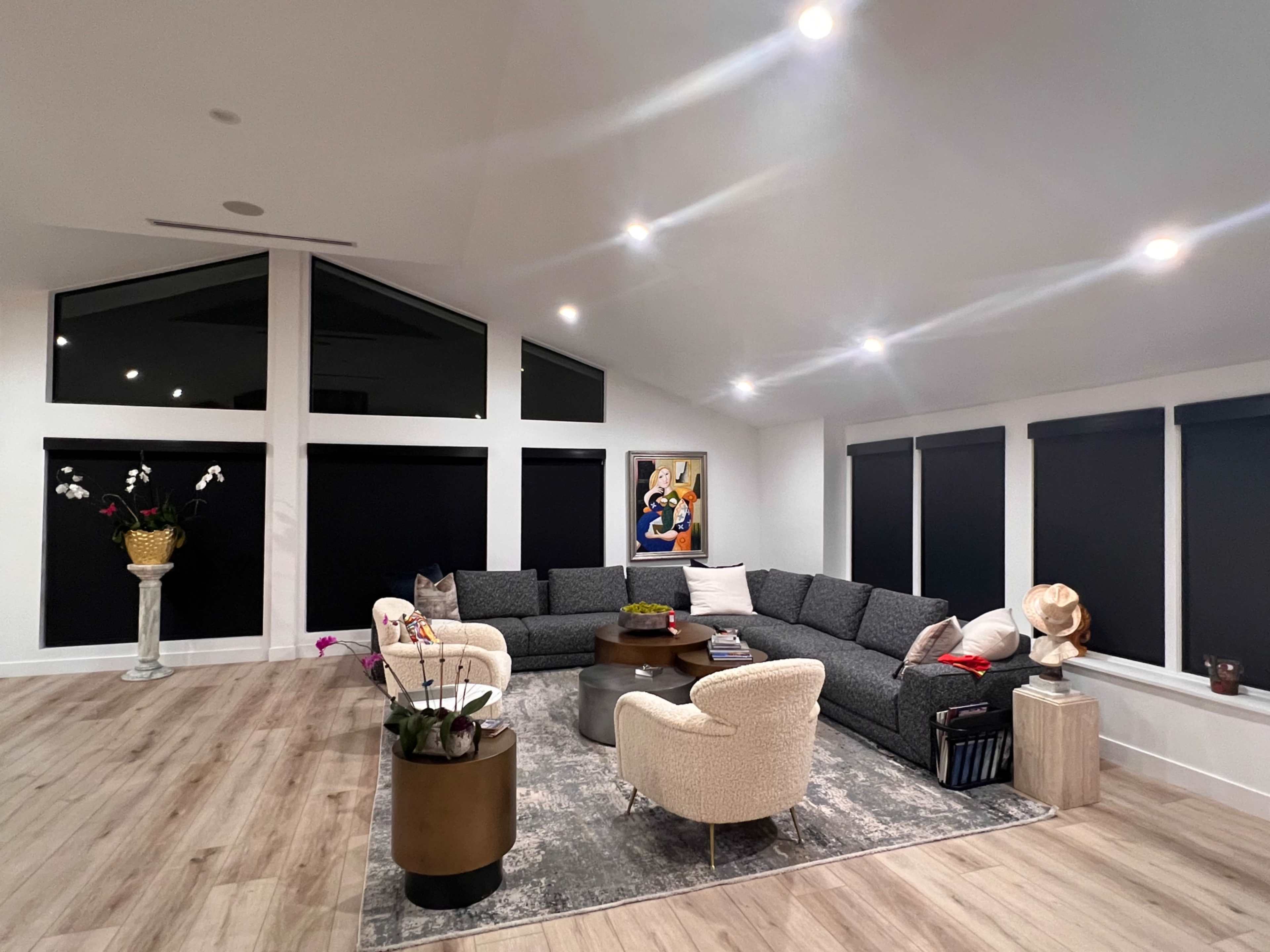The spacious living room features a large gray sectional couch, modern decor, and floor-to-ceiling windows with dark shades.