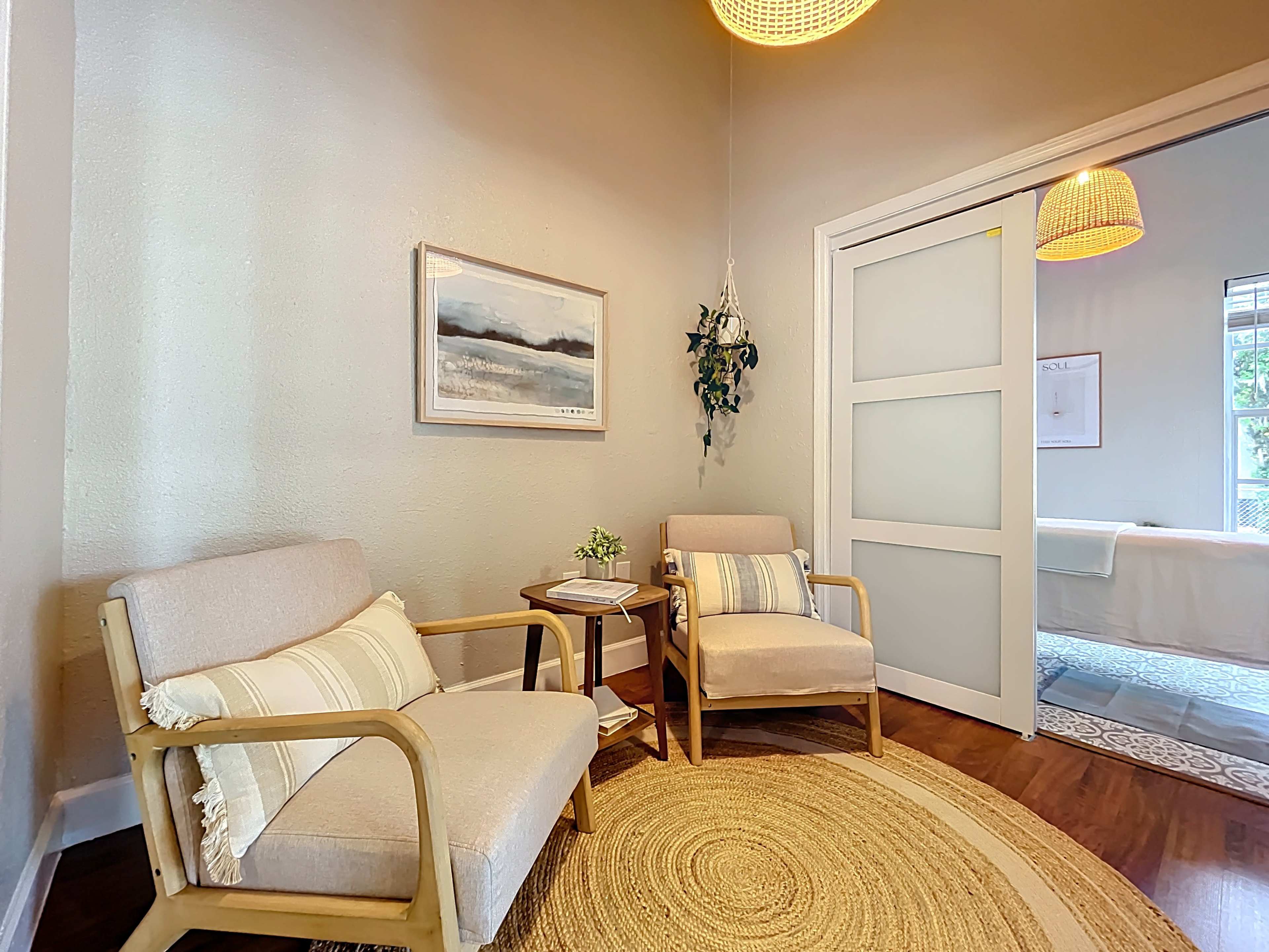 The image shows a small seating area with two chairs, a round rug, and a potted plant, accompanied by soft lighting and a framed landscape picture on the wall.