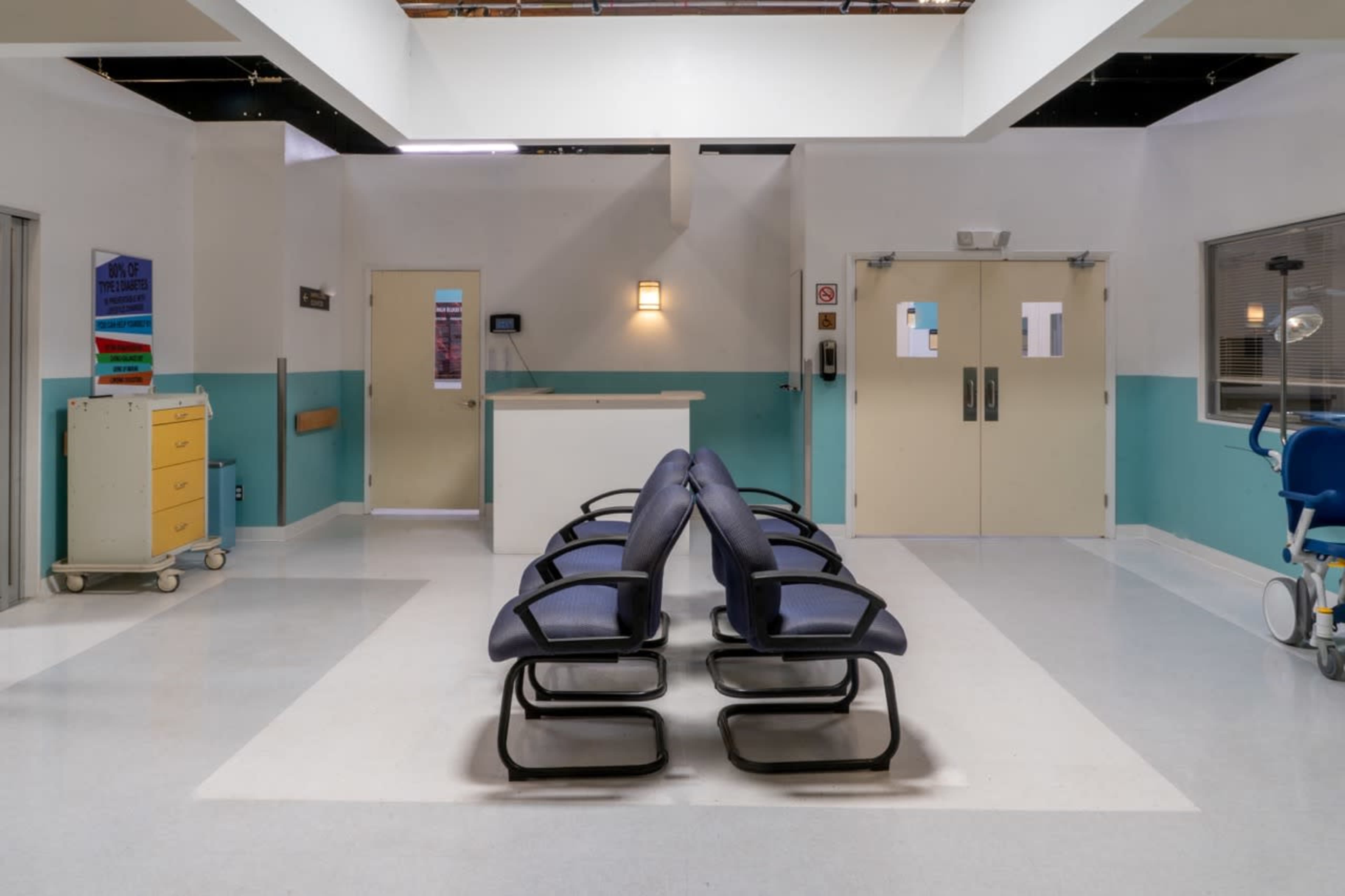 A waiting area in a medical facility with several black chairs arranged in a row, flanked by doors and a reception desk.