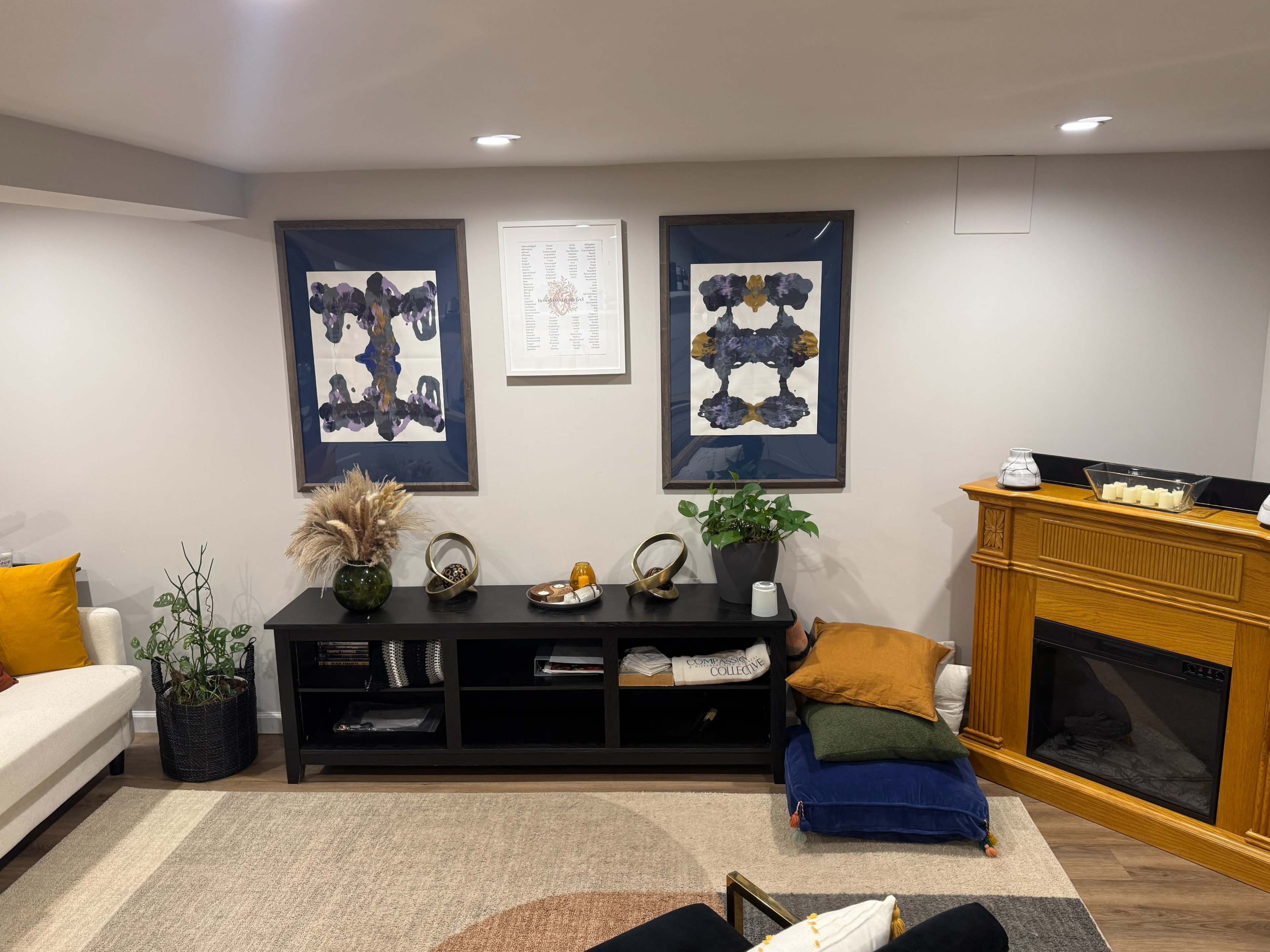 The image shows a cozy living room with two framed artworks on the wall, a black shelf with decorative items, and a small seating area featuring cushions and a fireplace.