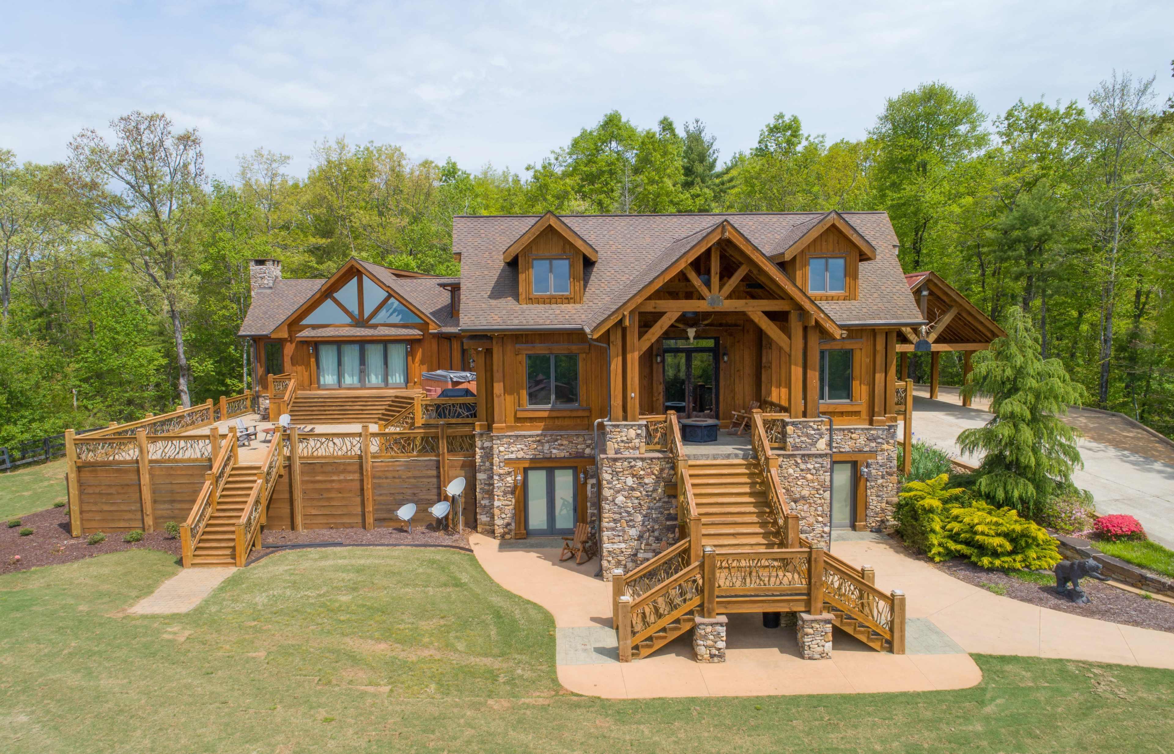 A large, wooden lodge with multiple levels, a stone foundation, and a spacious deck surrounded by greenery.