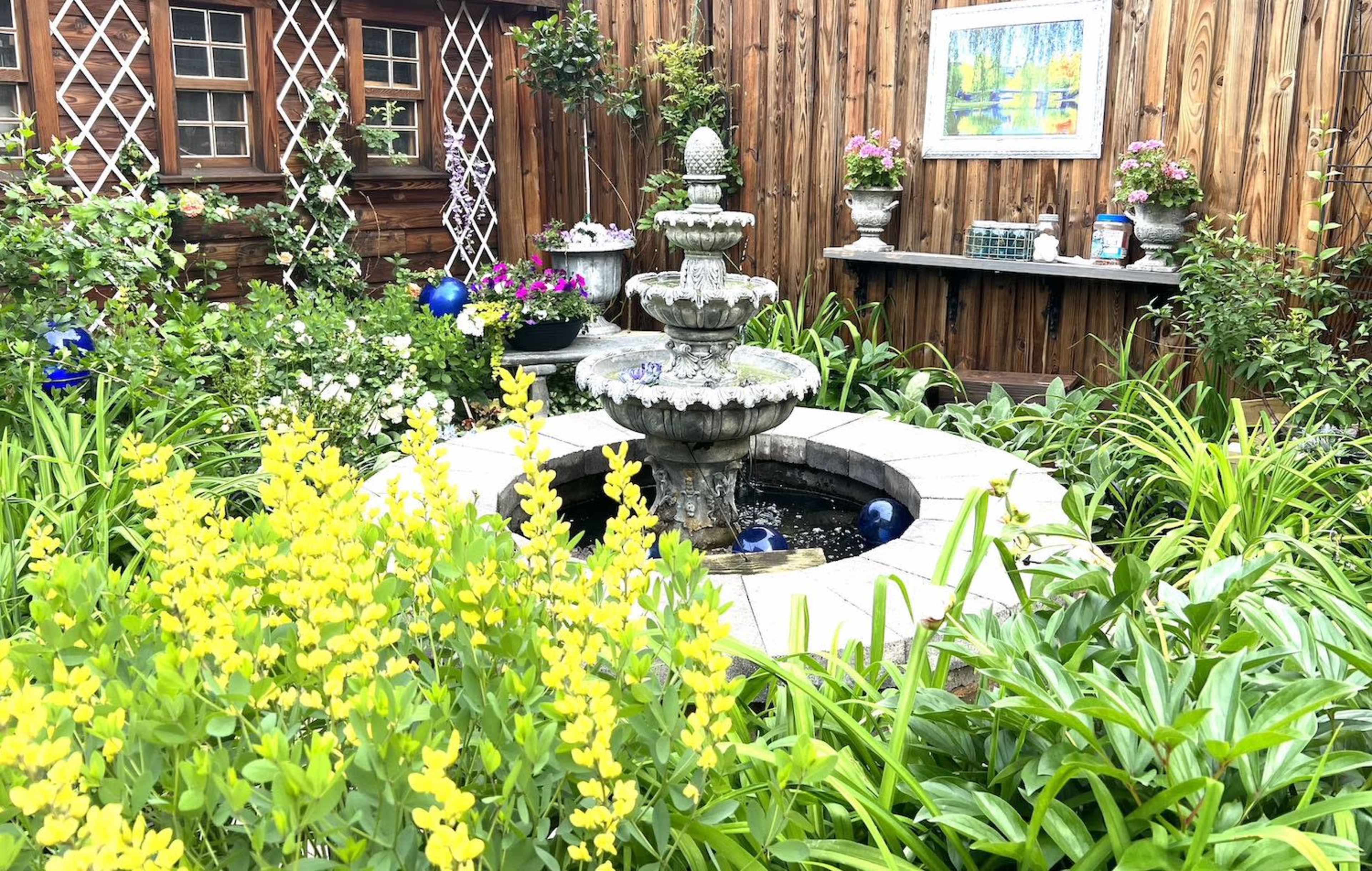 A stone fountain stands in the center of a landscaped garden filled with flowers and greenery, surrounded by a wooden fence and decorative elements.