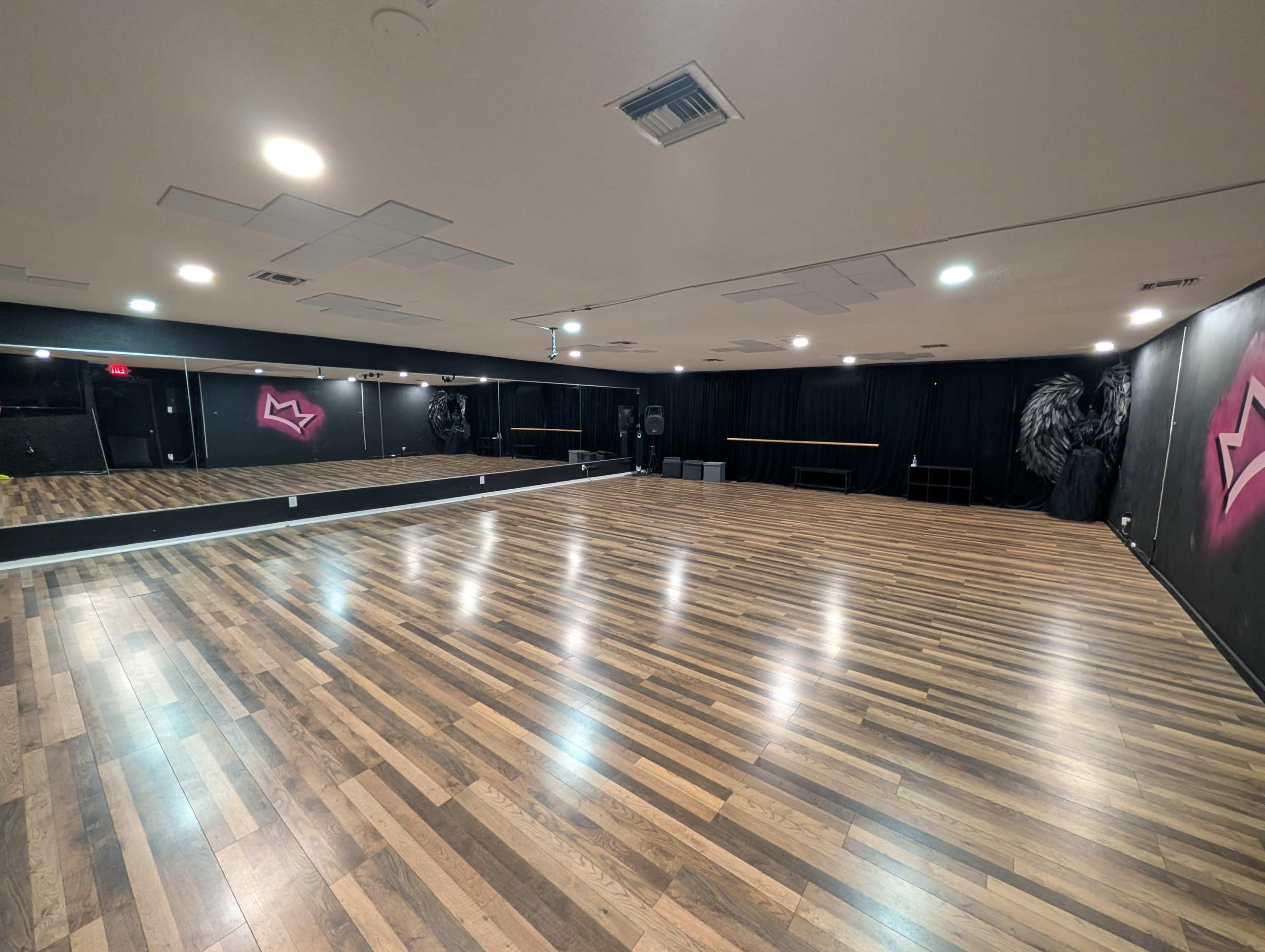 A spacious dance studio features a polished wooden floor, mirrors along one wall, and black curtains with a mural on another wall.