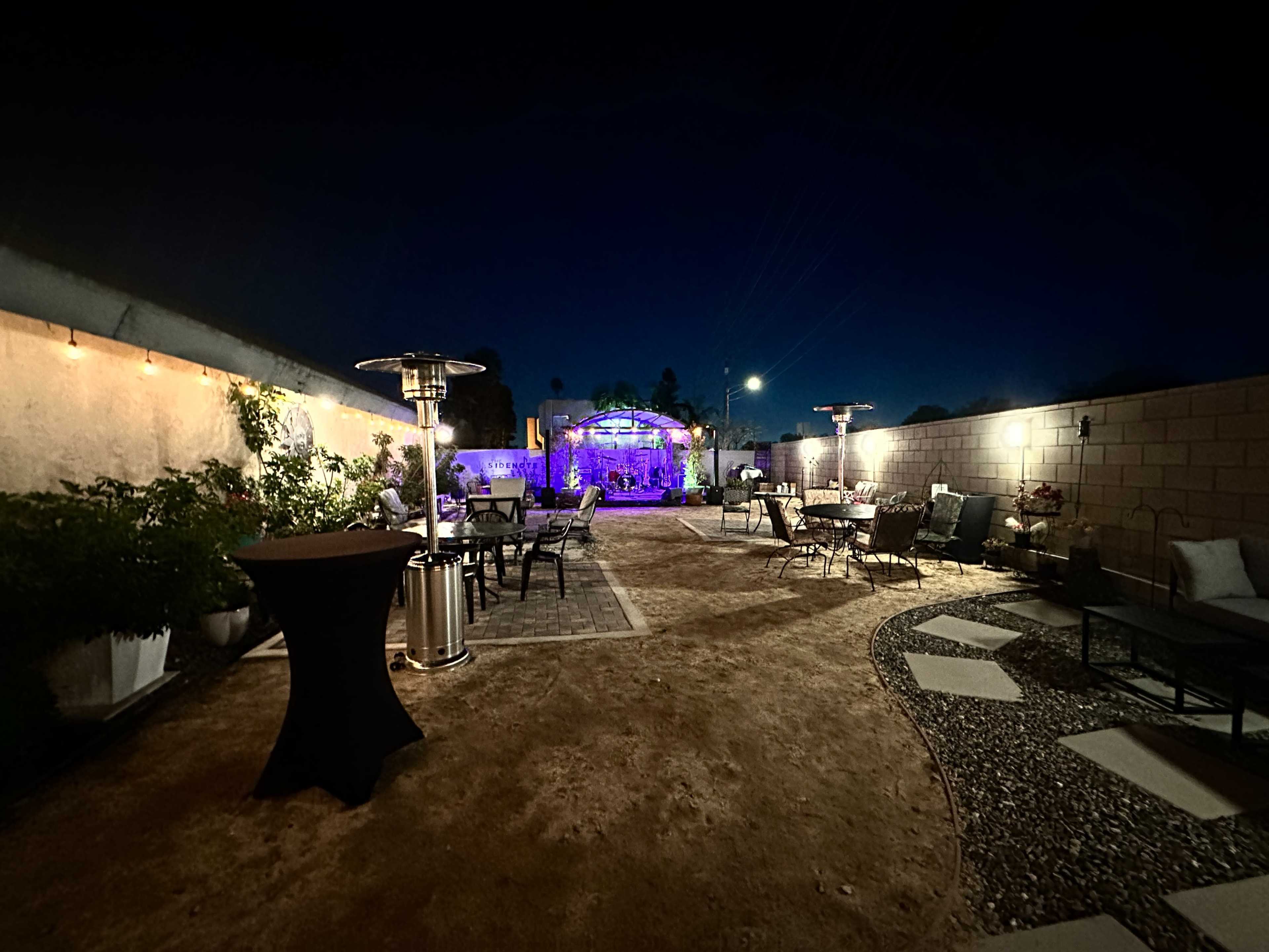 The image shows a nighttime outdoor patio area with seating, decorative lighting, and a pathway leading to a lit structure in the background.