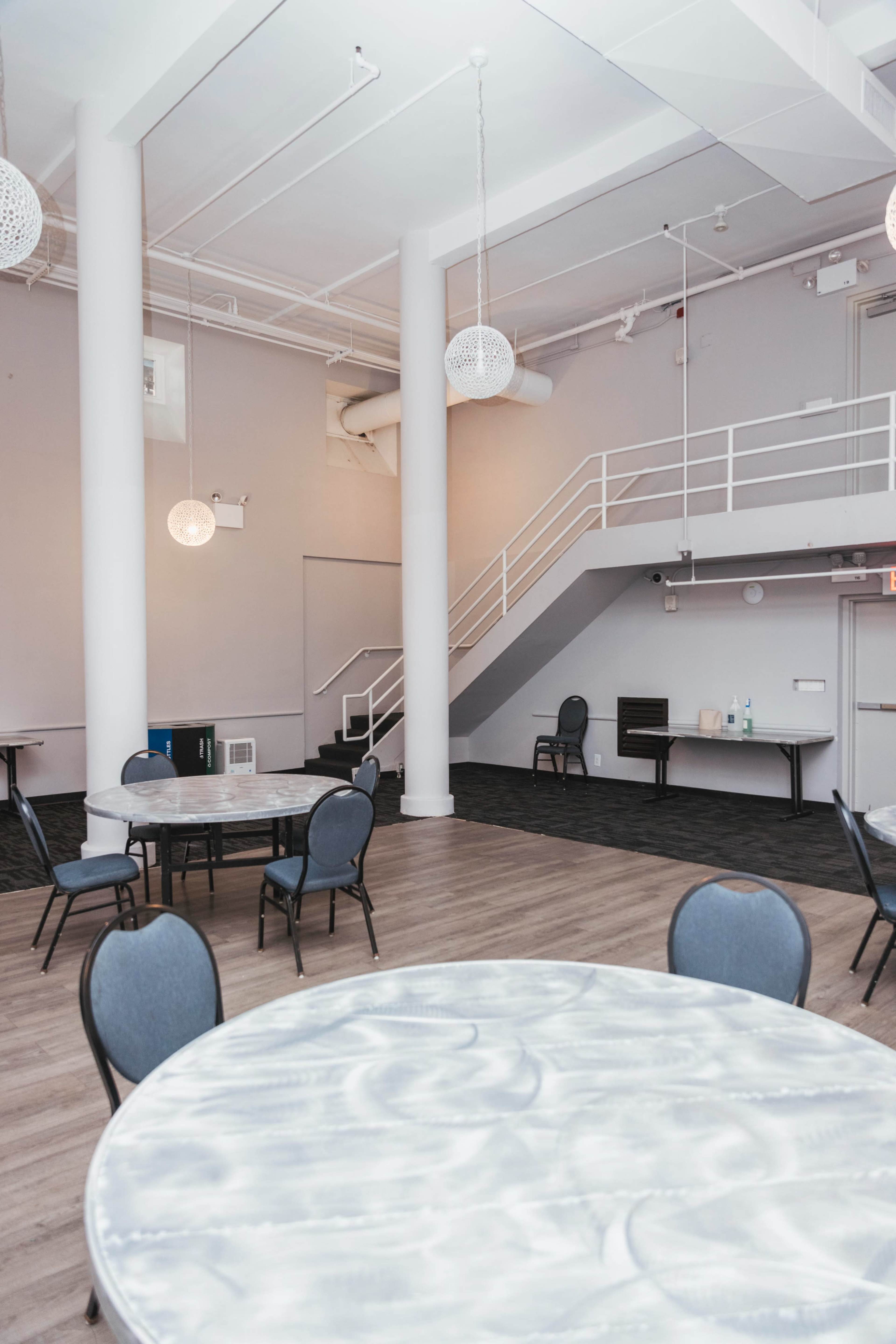 The image shows a spacious event room with several round tables and chairs, featuring a modern staircase and pendant lighting.