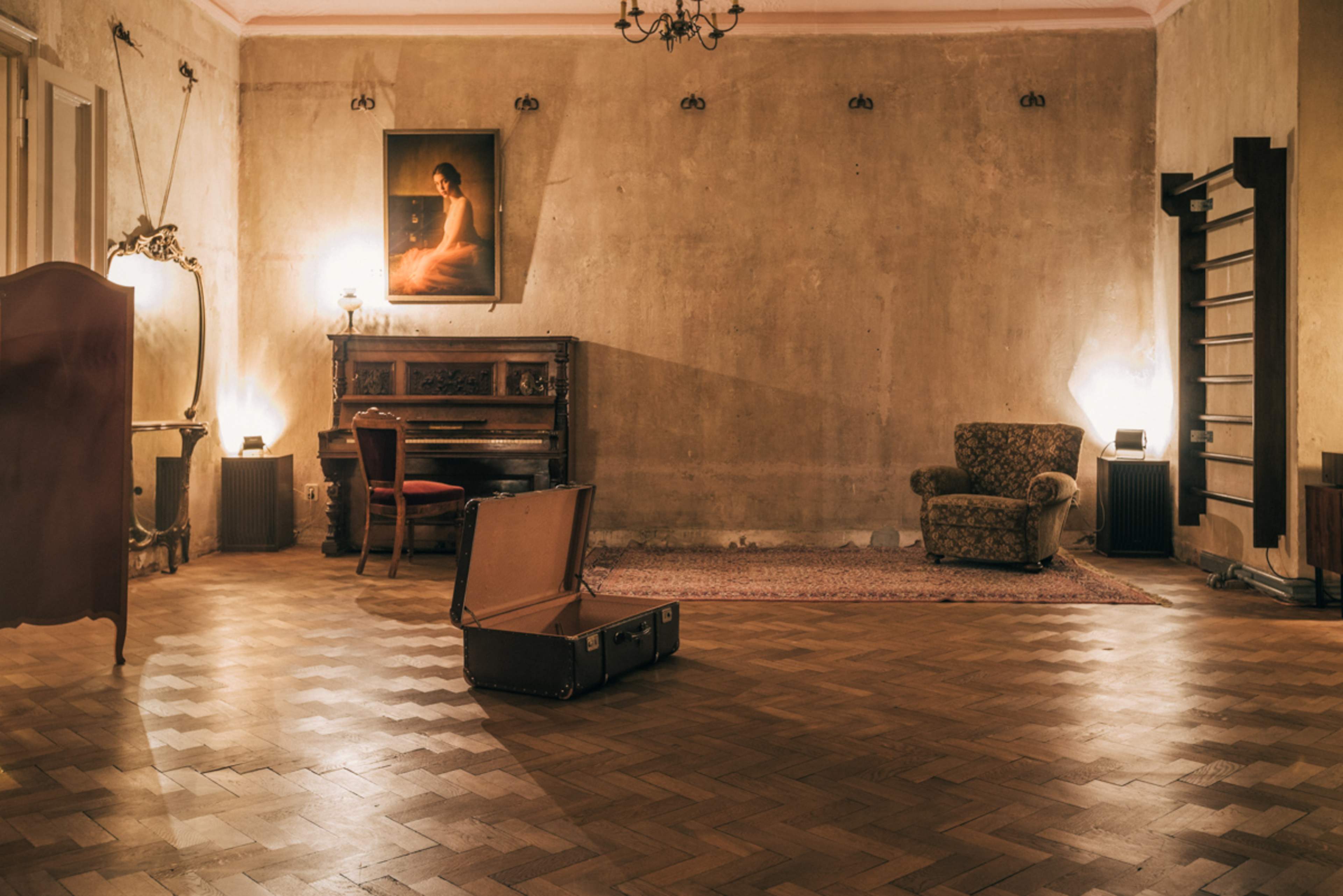 A vintage suitcase lies open on the wooden floor of a dimly lit, empty room featuring a piano, a patterned armchair, and a portrait hanging on the wall.