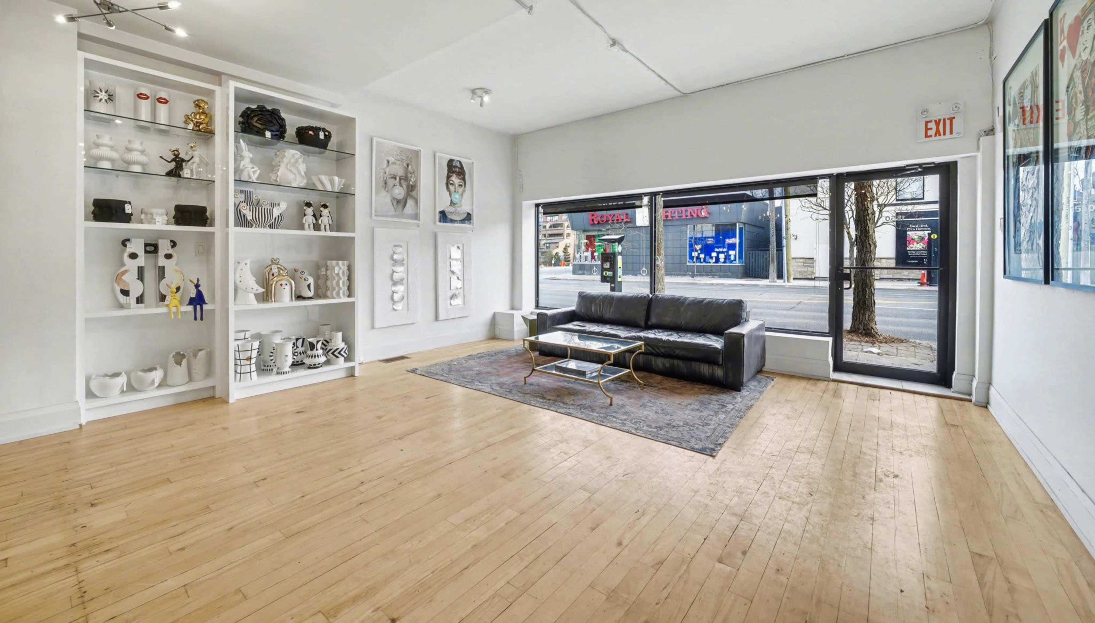 The image shows a modern, minimalist interior room with a wooden floor, featuring a black sofa, a coffee table, and a bookshelf filled with decorative objects, all illuminated by natural light coming through large windows.