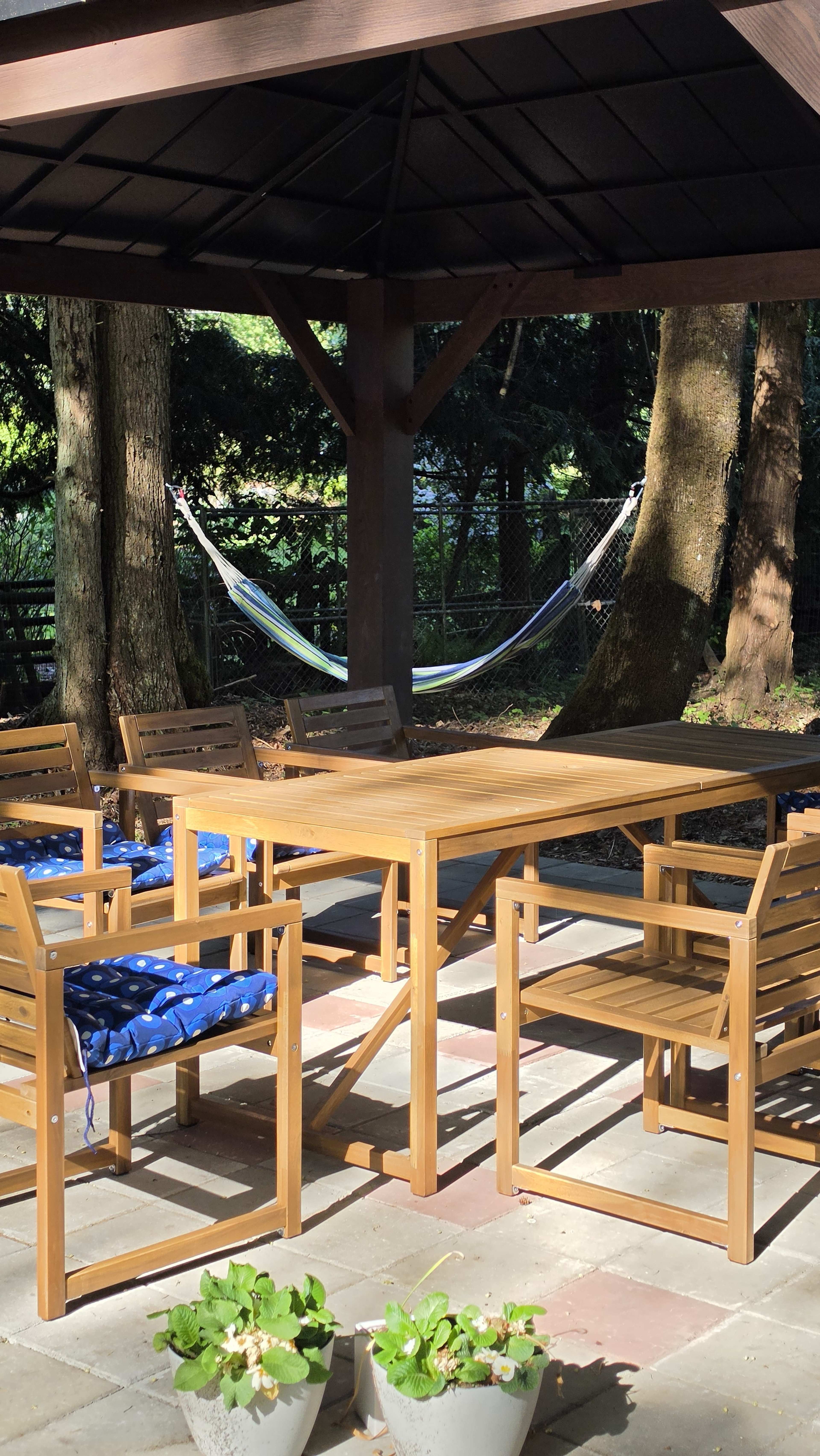 A wooden dining table with chairs sits under a gazebo, accompanied by a hammock strung between two trees in the background.