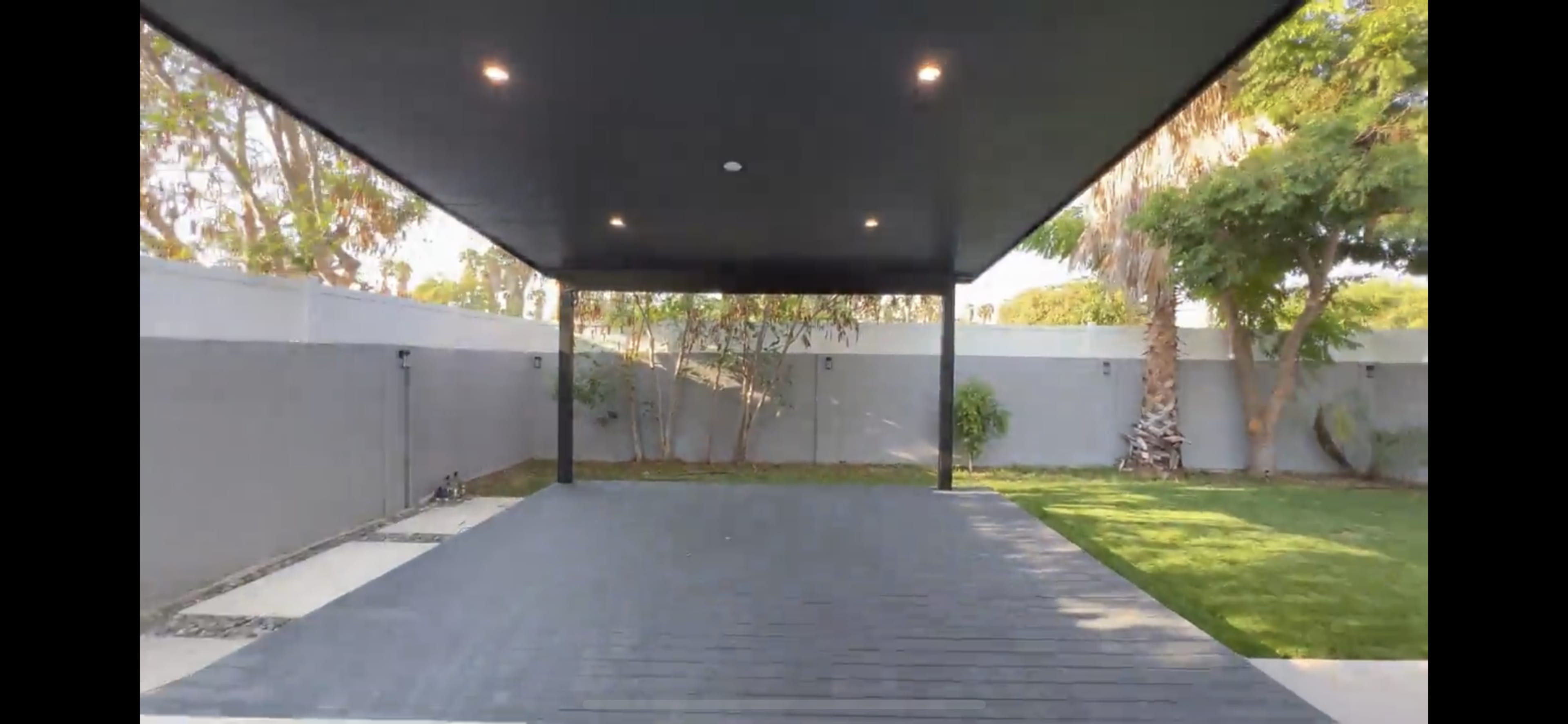 A covered outdoor patio area with a wooden deck and a lawn surrounded by gray walls and greenery.
