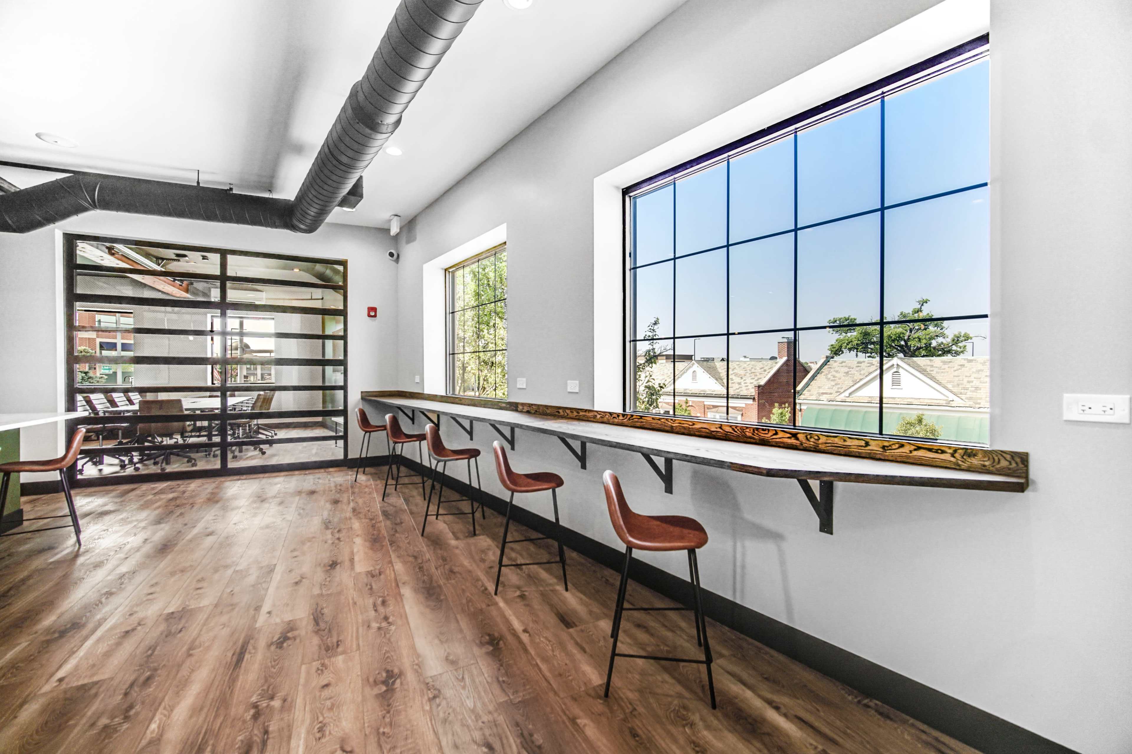 A modern interior space with a long wooden counter and several high stools positioned near large windows overlooking a neighborhood.
