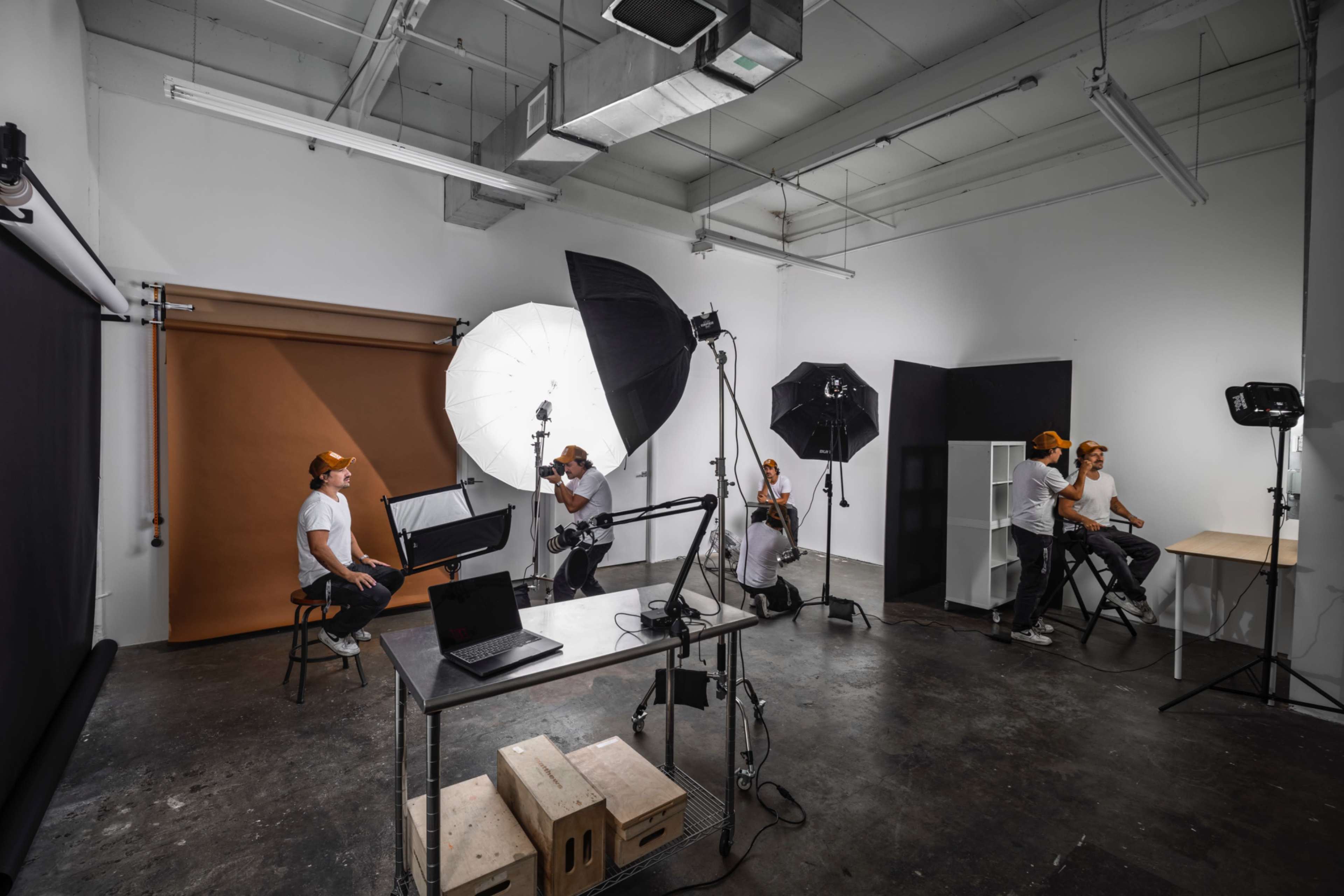 A photography studio is set up with several lighting equipment and multiple photographers capturing a model seated on a stool.