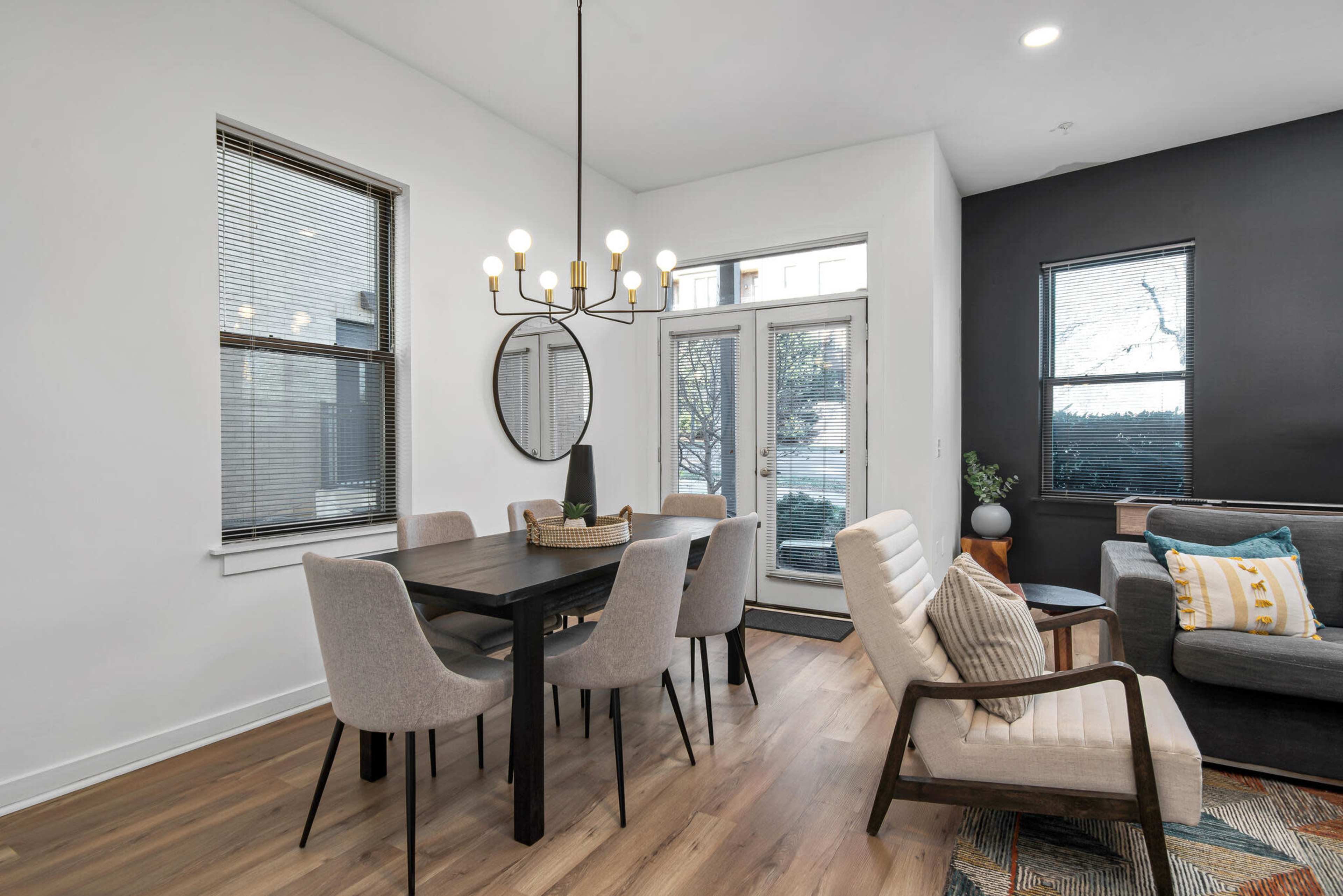 The image shows a modern dining area with a black table surrounded by beige chairs, a decorative mirror on the wall, and large windows allowing natural light.
