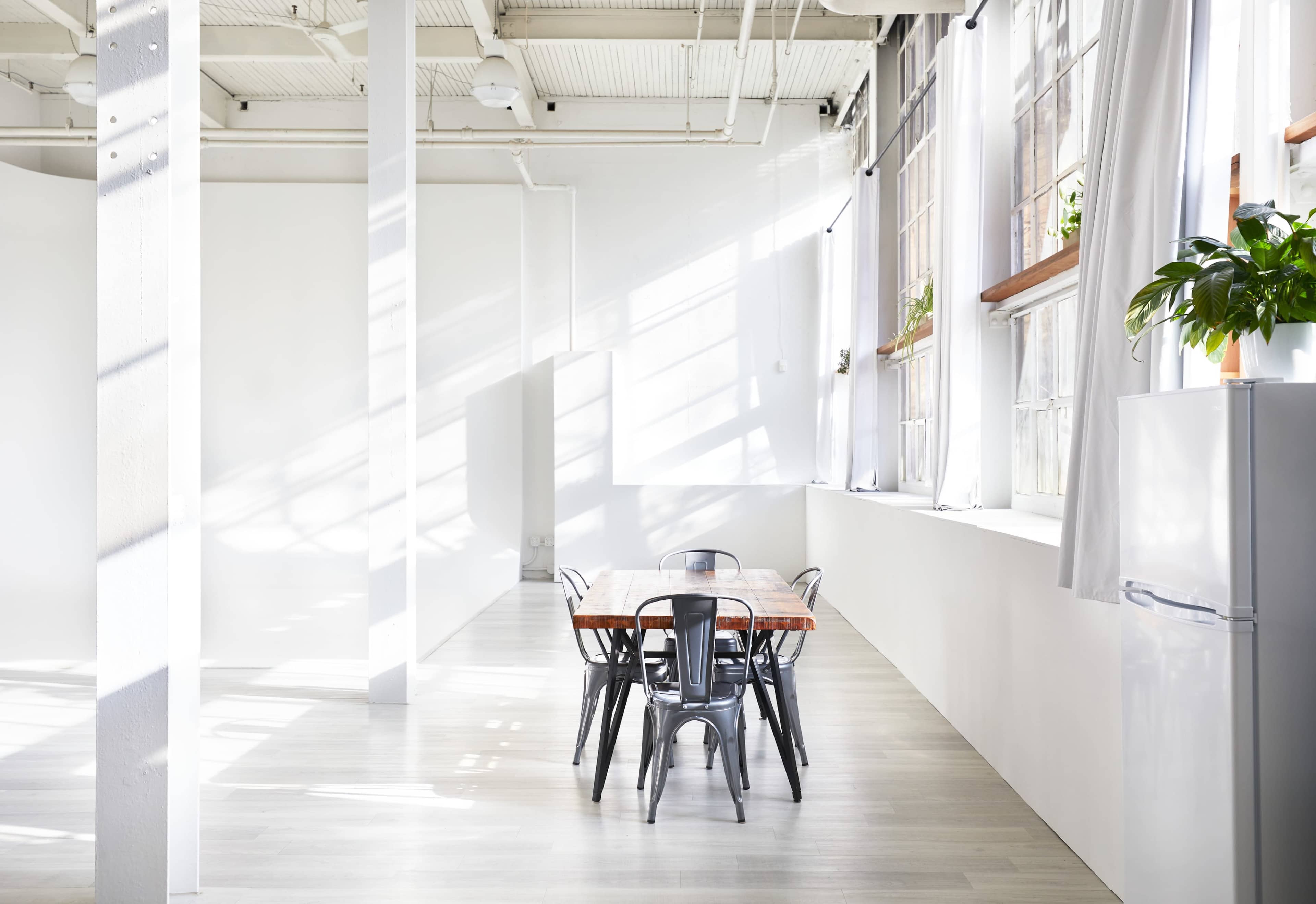The image shows a bright, spacious room with large windows, a wooden table surrounded by black chairs, and a refrigerator in the corner.