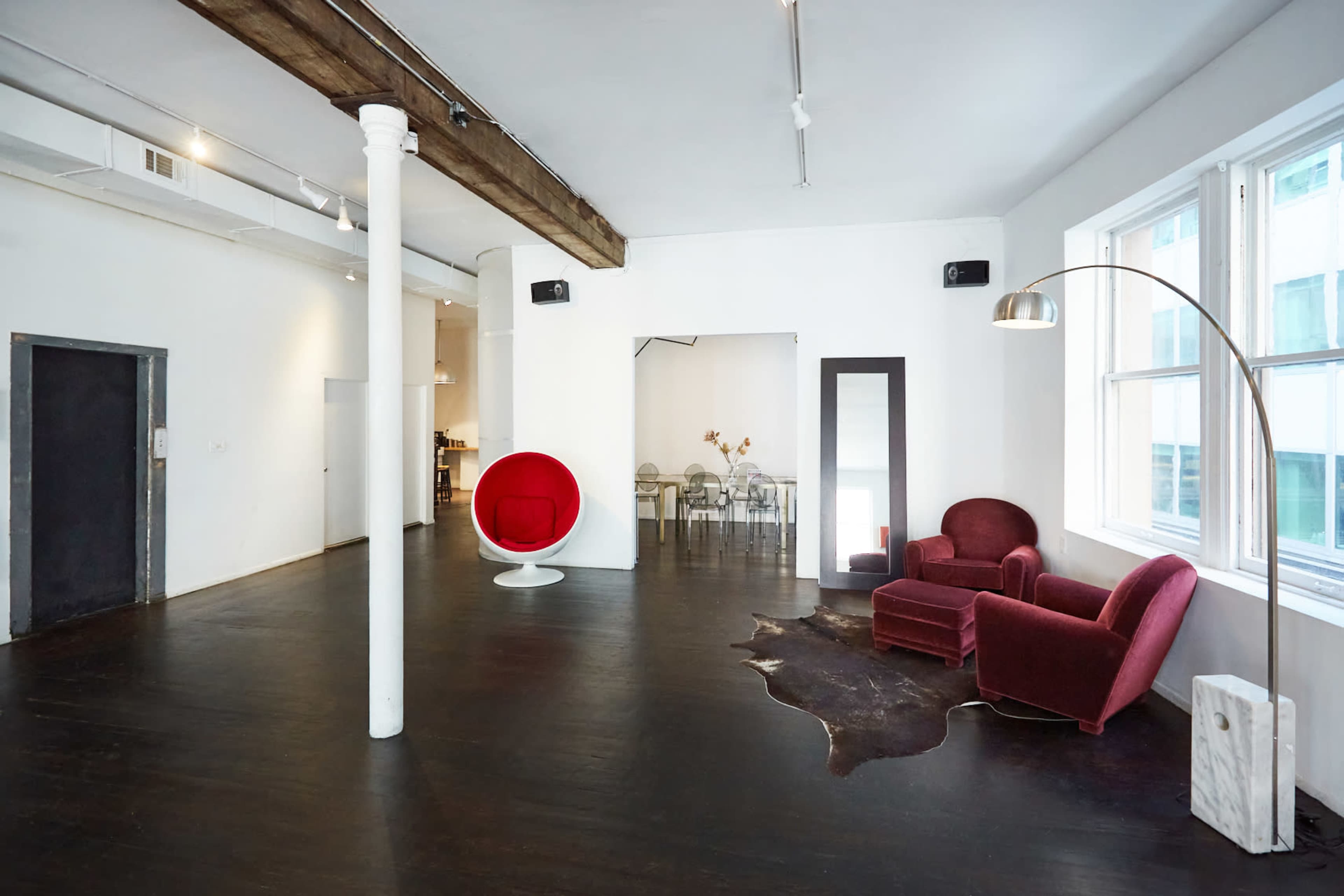 A spacious, modern interior with a wooden beam ceiling, featuring a red chair and a white round chair, a mirror, and a dining area in the background.