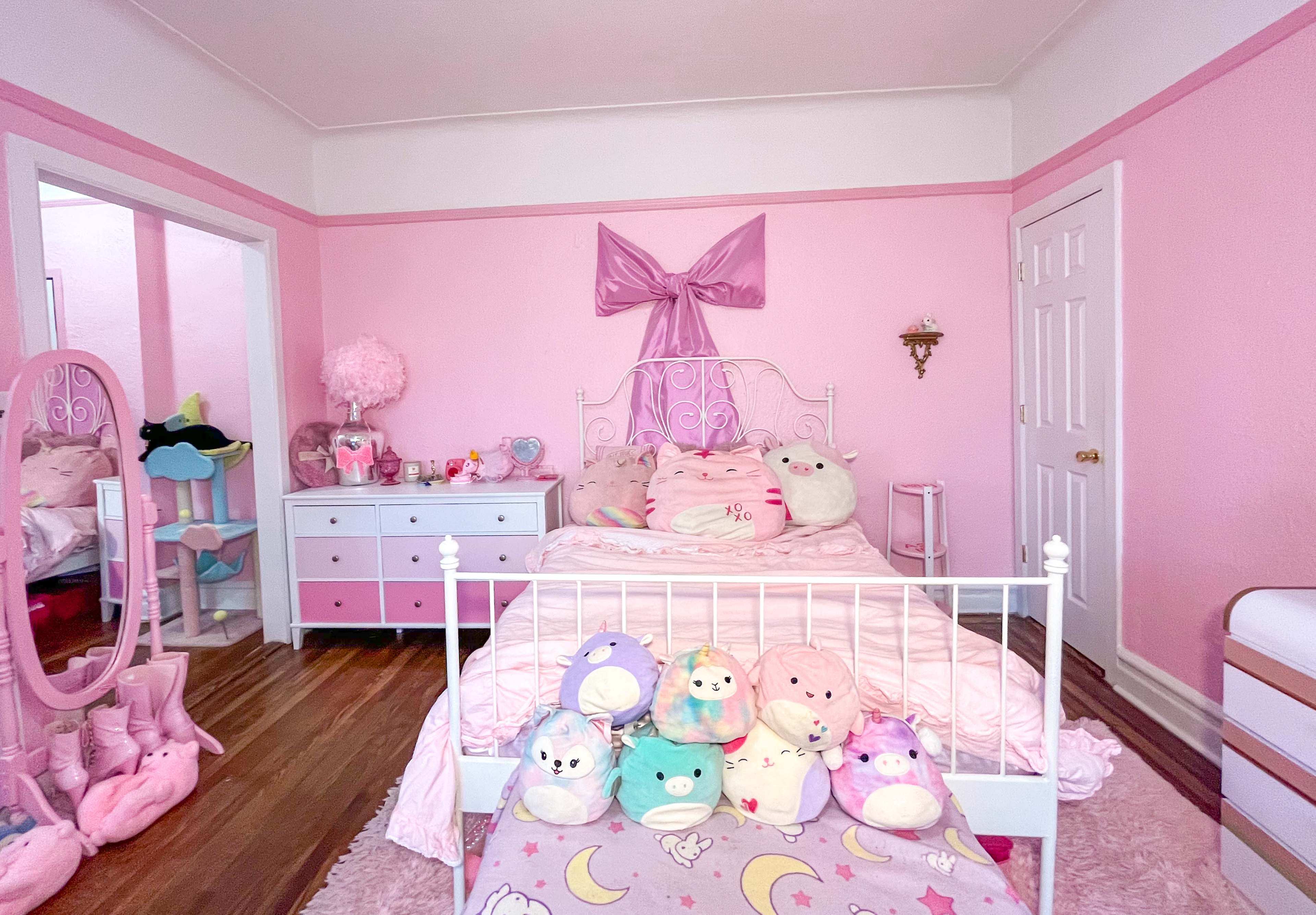 A pink-themed bedroom with a bed adorned with plush toys, a dresser, and a mirror.