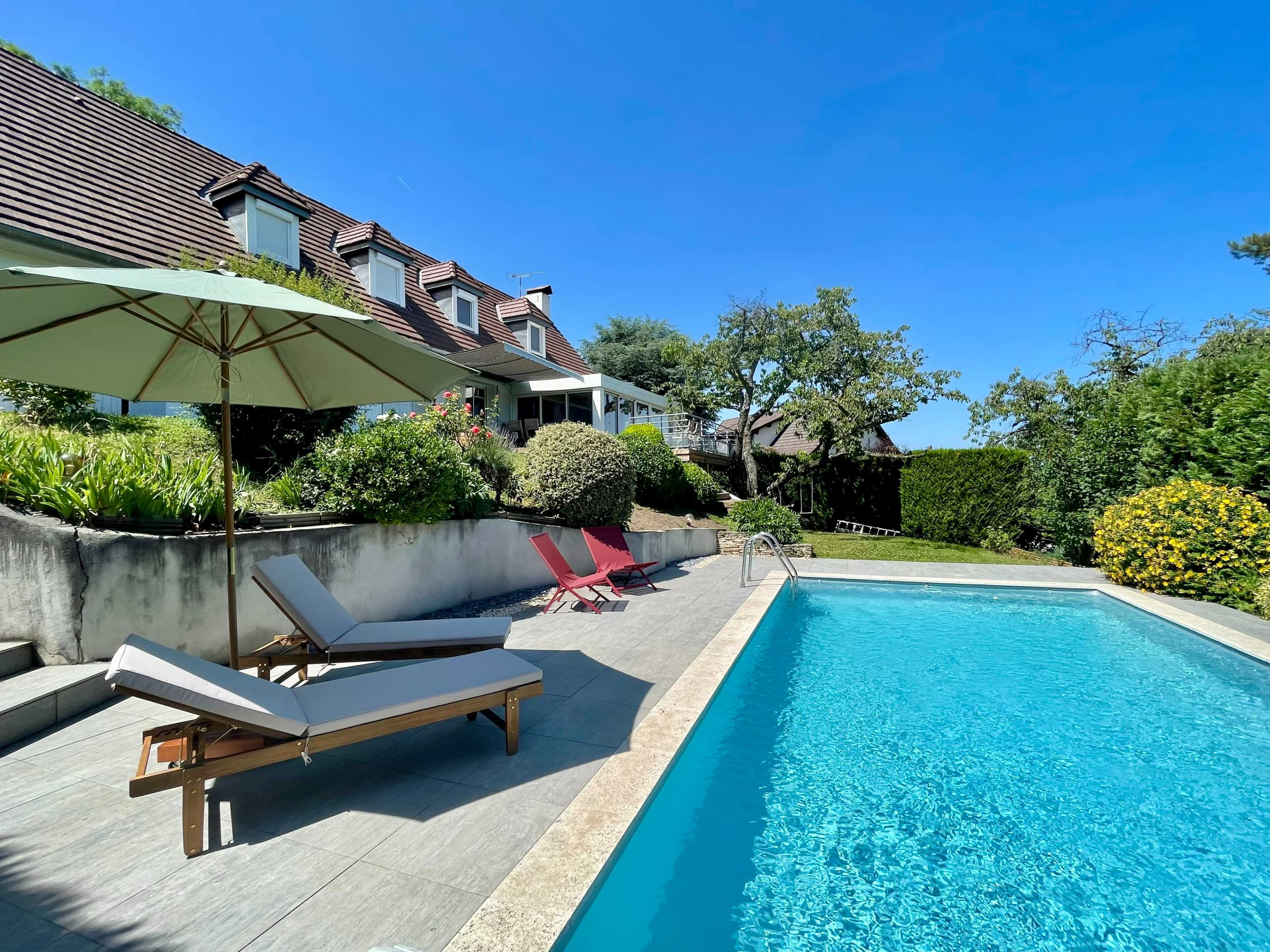 The image shows a swimming pool bordered by lounge chairs and an umbrella in a backyard garden with a house and trees in the background.