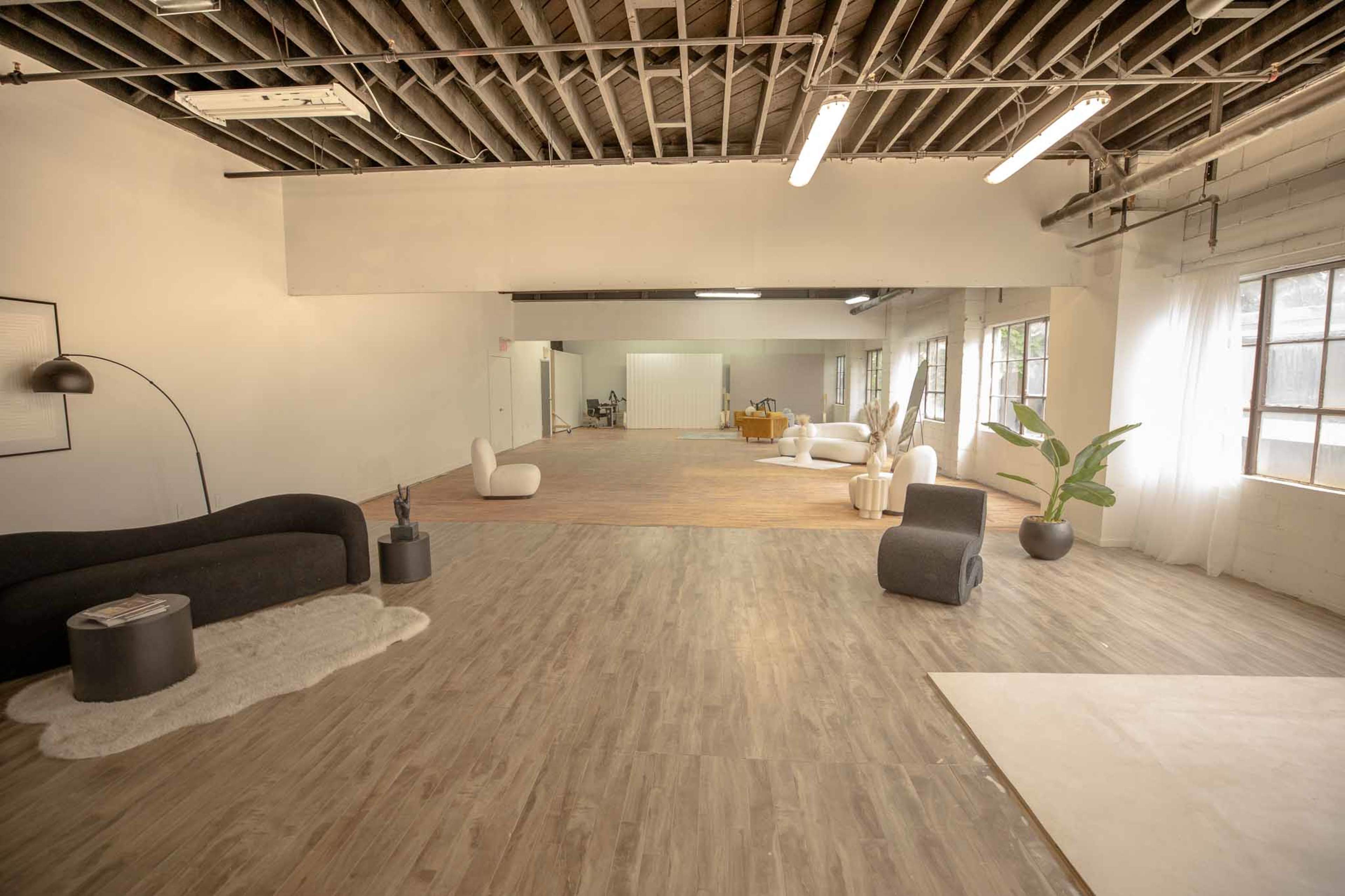 The image shows a spacious, modern studio with hardwood floors, large windows, and various seating arrangements, including a black couch and a plant.