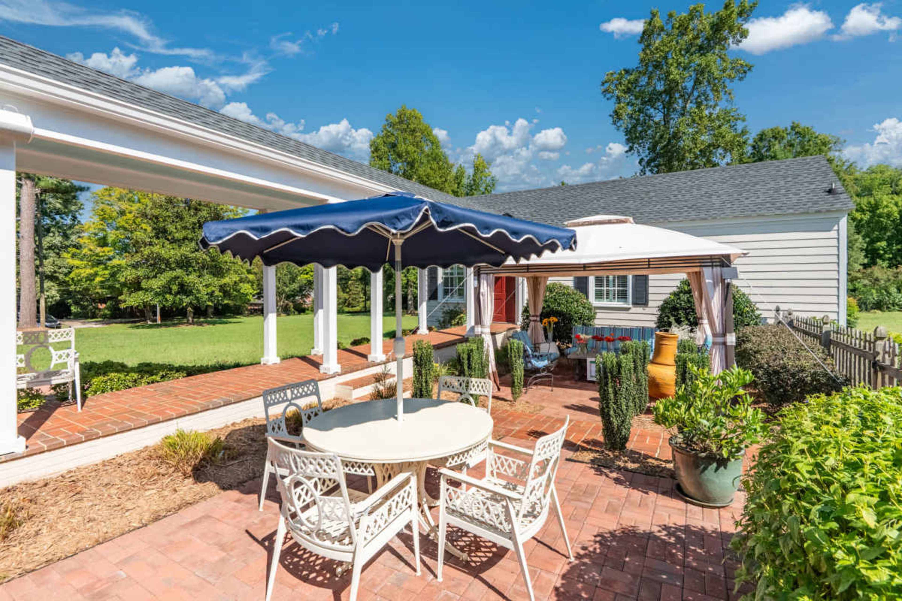The image shows a patio area with a round table, chairs, and two umbrellas, surrounded by greenery and a single-story house in the background.