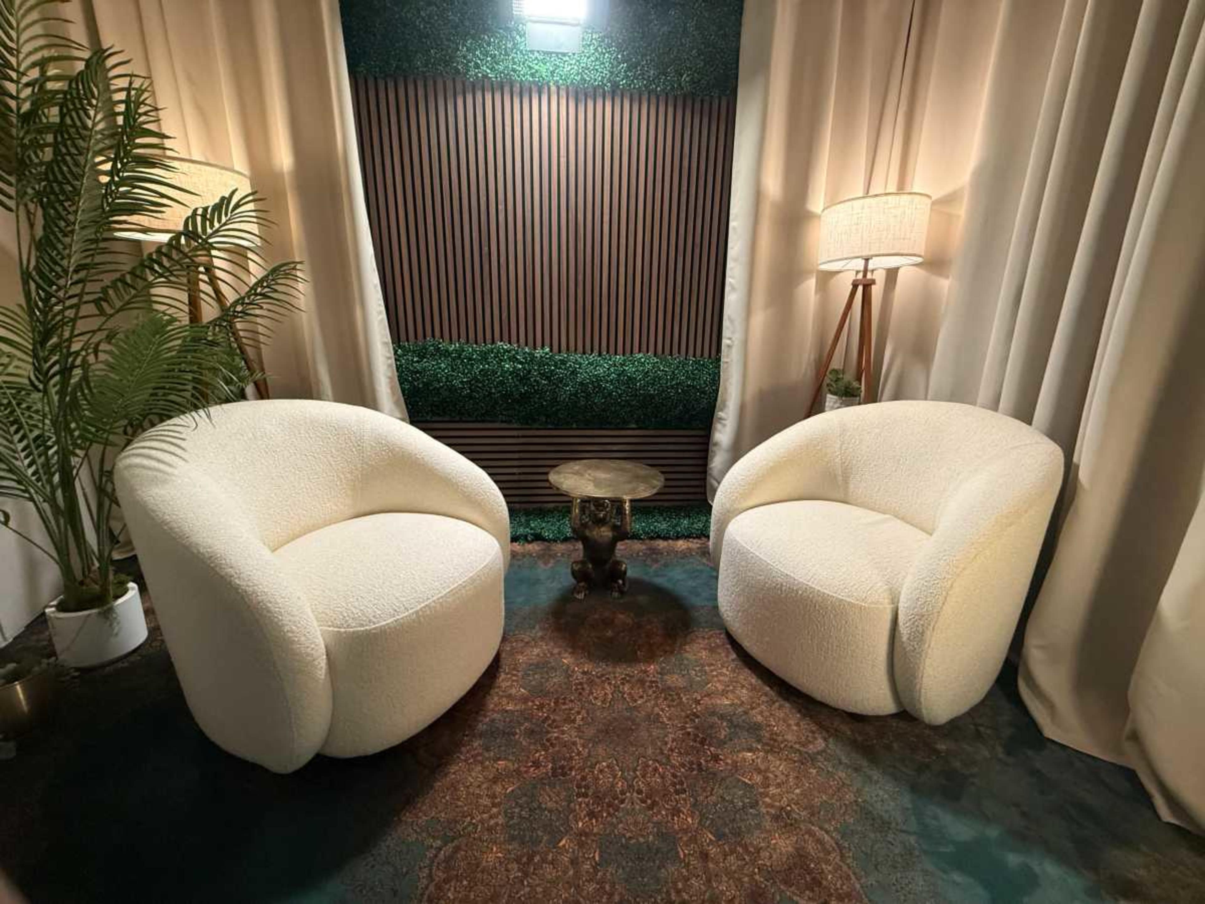 The image shows two white, rounded armchairs facing each other on a patterned rug, with a small table between them and decorative plants in the background.
