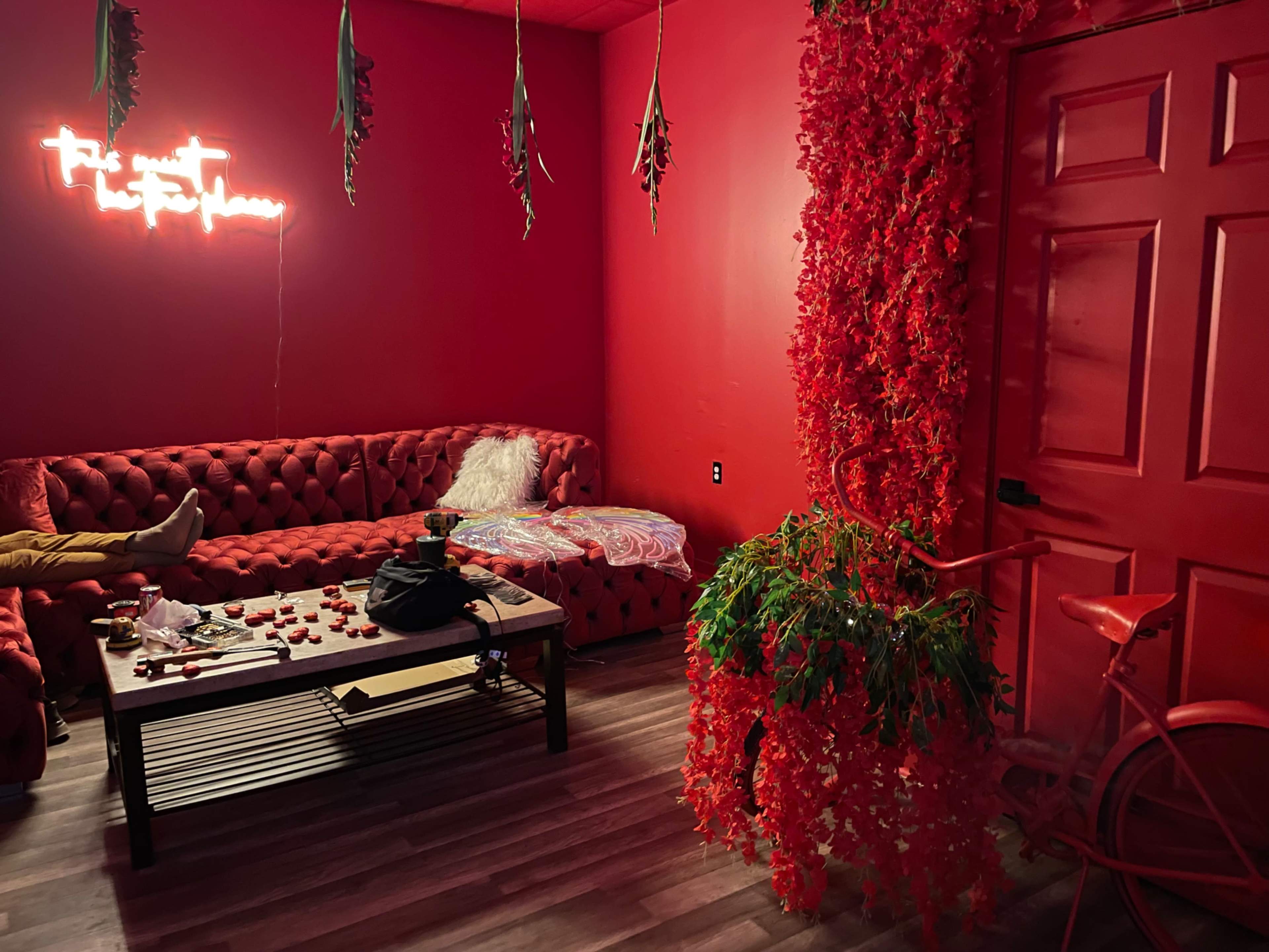 The room features red walls, a red tufted couch, a coffee table scattered with rose petals, and a bicycle adorned with artificial flowers at the entrance.