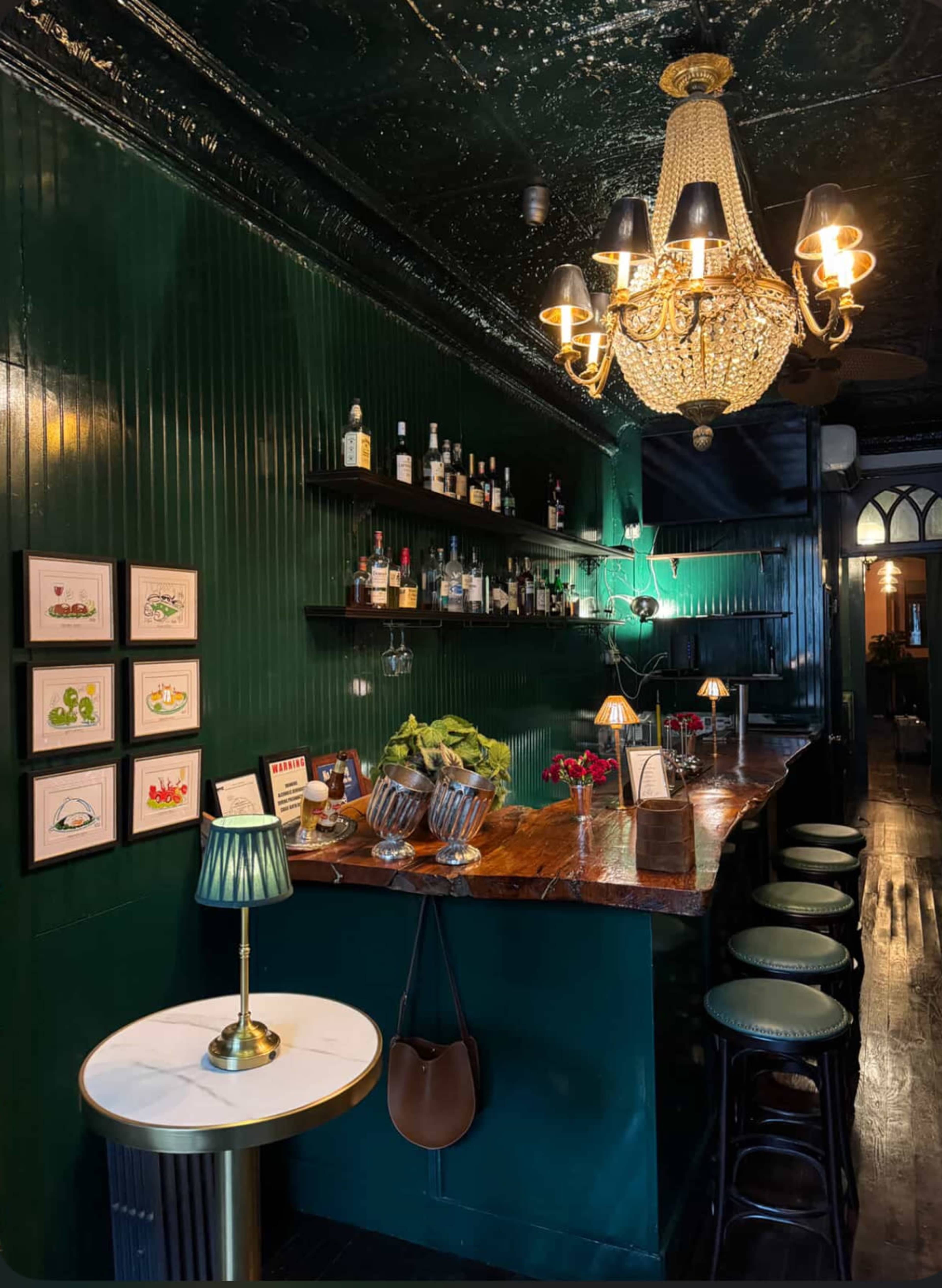 The image shows an elegant bar with a dark green wall, a polished wooden counter, and a stylish chandelier, featuring shelves of various liquor bottles.