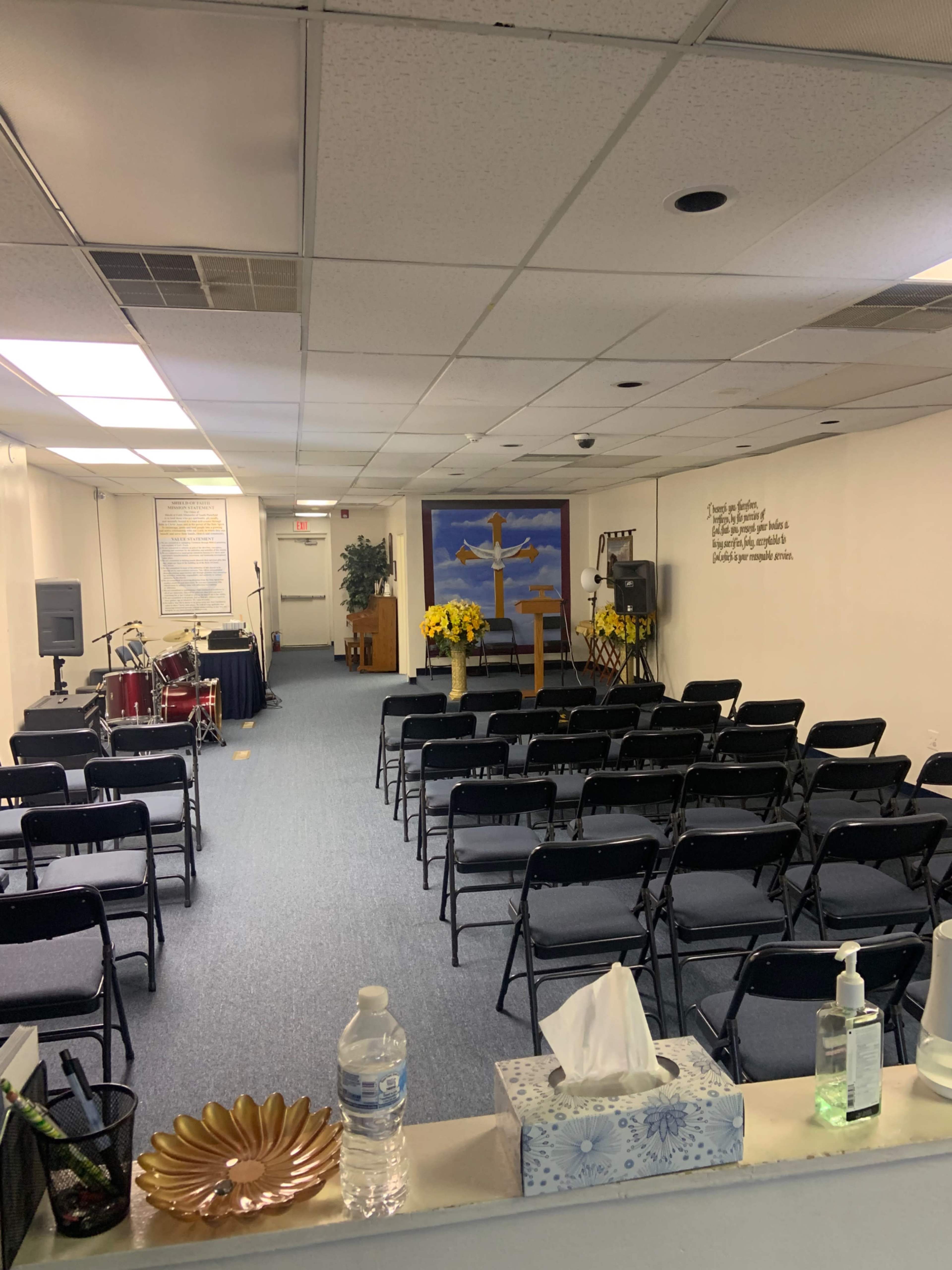 A modest indoor space set up for a gathering, featuring rows of black chairs facing a stage with a microphone, drums, and a backdrop displaying a cross and a scenic painting.