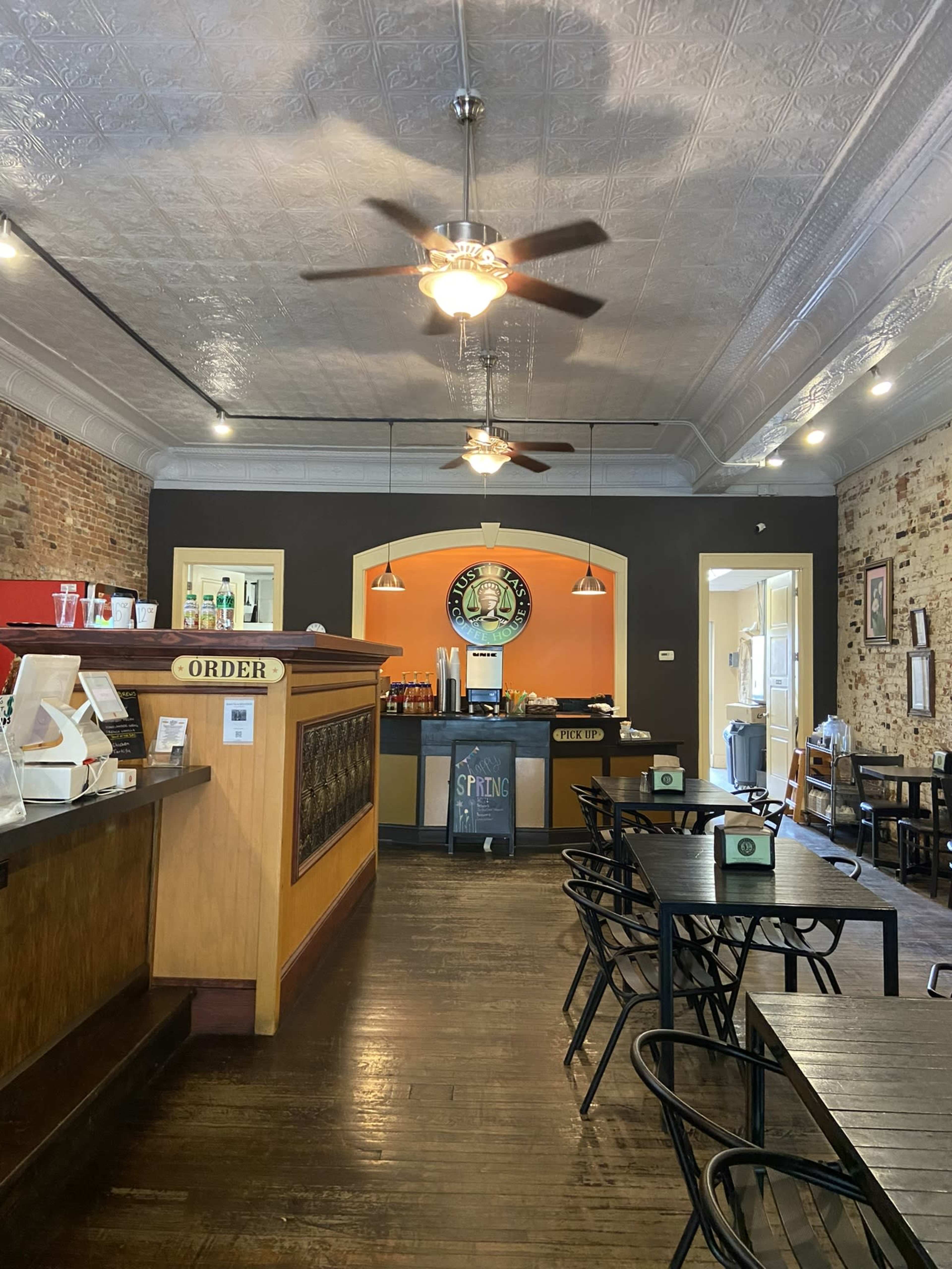 The image shows a spacious cafe with wooden tables and chairs, a counter with an "Order" sign, and decorative ceiling fans above.