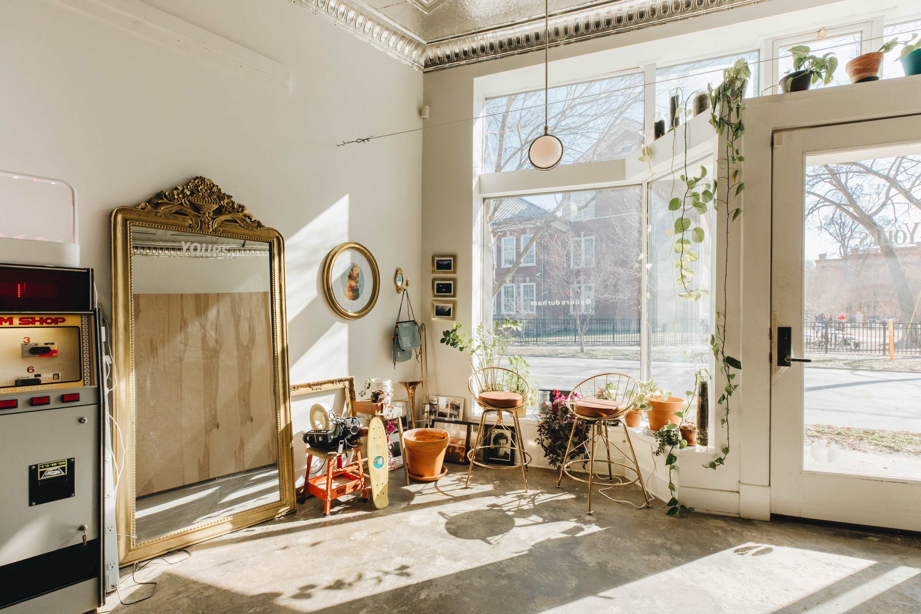 A cozy interior space with a large ornate mirror, several potted plants, and two rattan chairs beside a bright window.