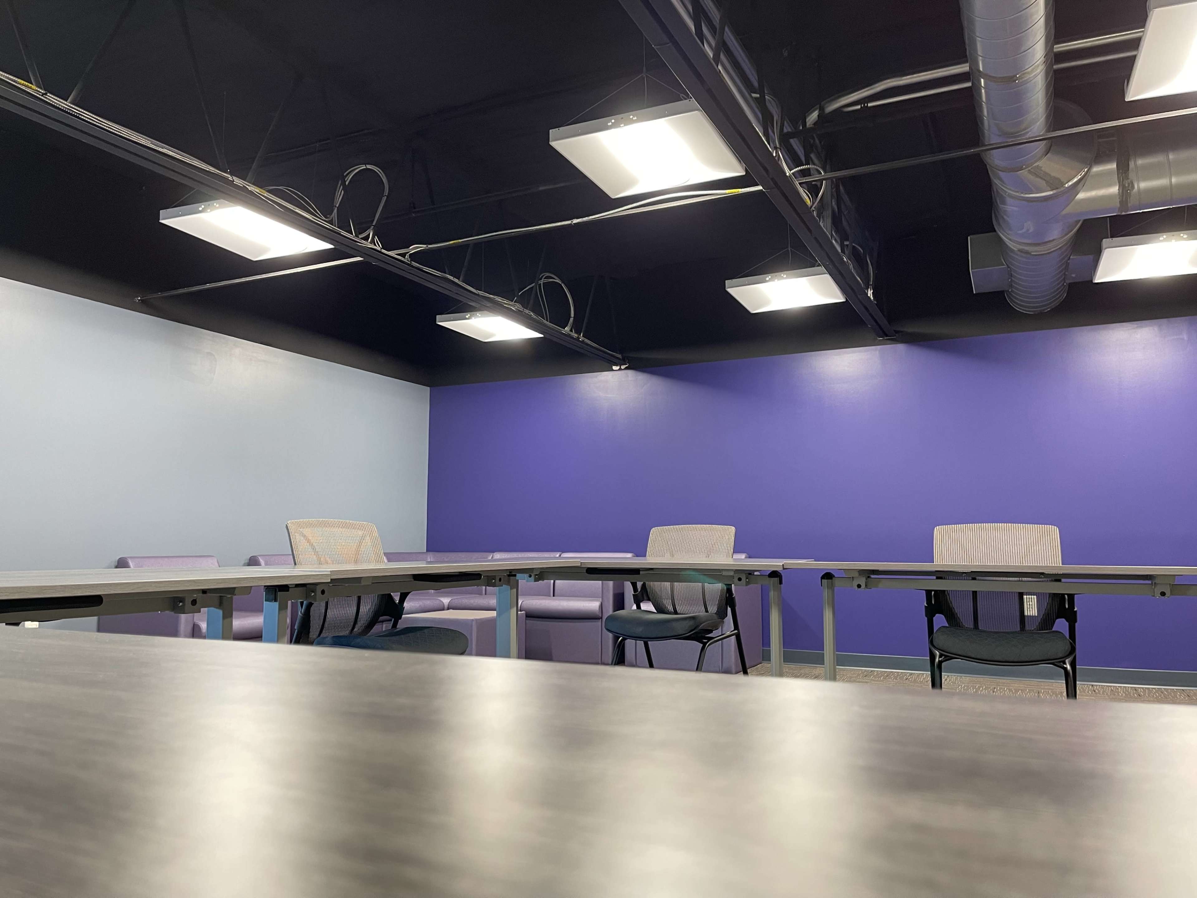 A conference room with several tables and chairs arranged in a U-shape, featuring a purple accent wall and overhead lighting.