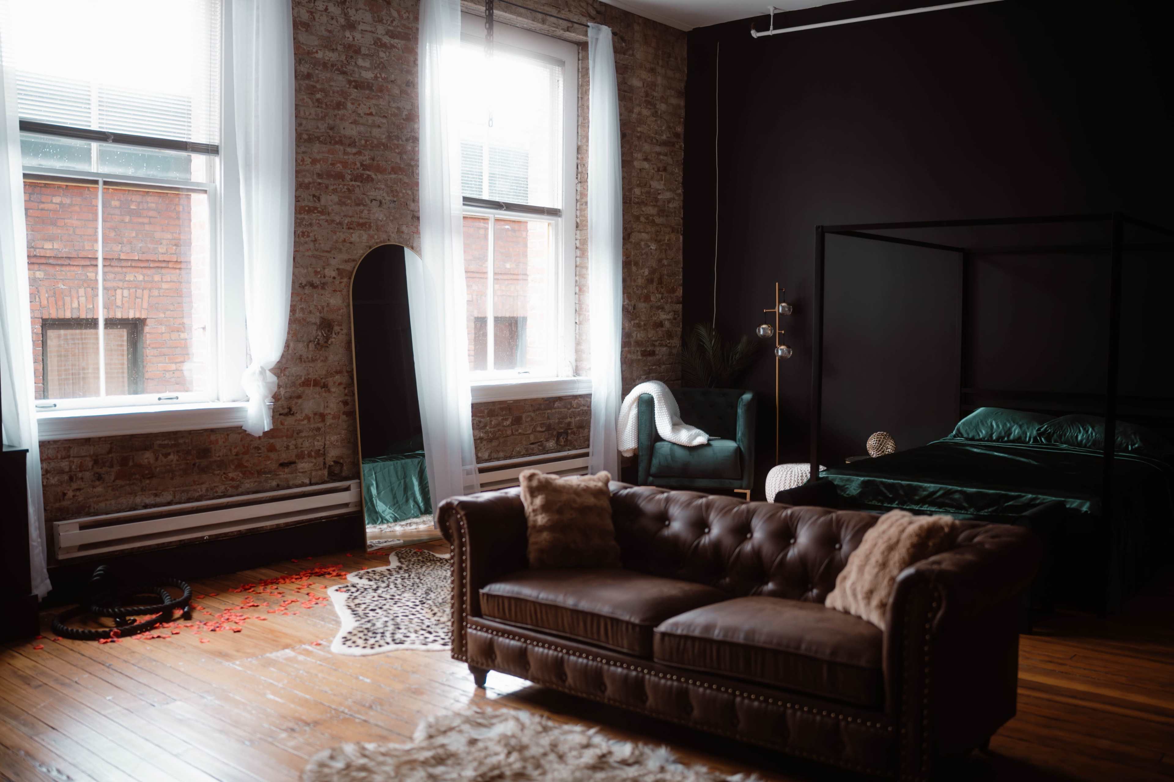 The image shows a modern bedroom with a four-poster bed, a large mirror, a green armchair, and a brown leather couch arranged on wooden flooring.
