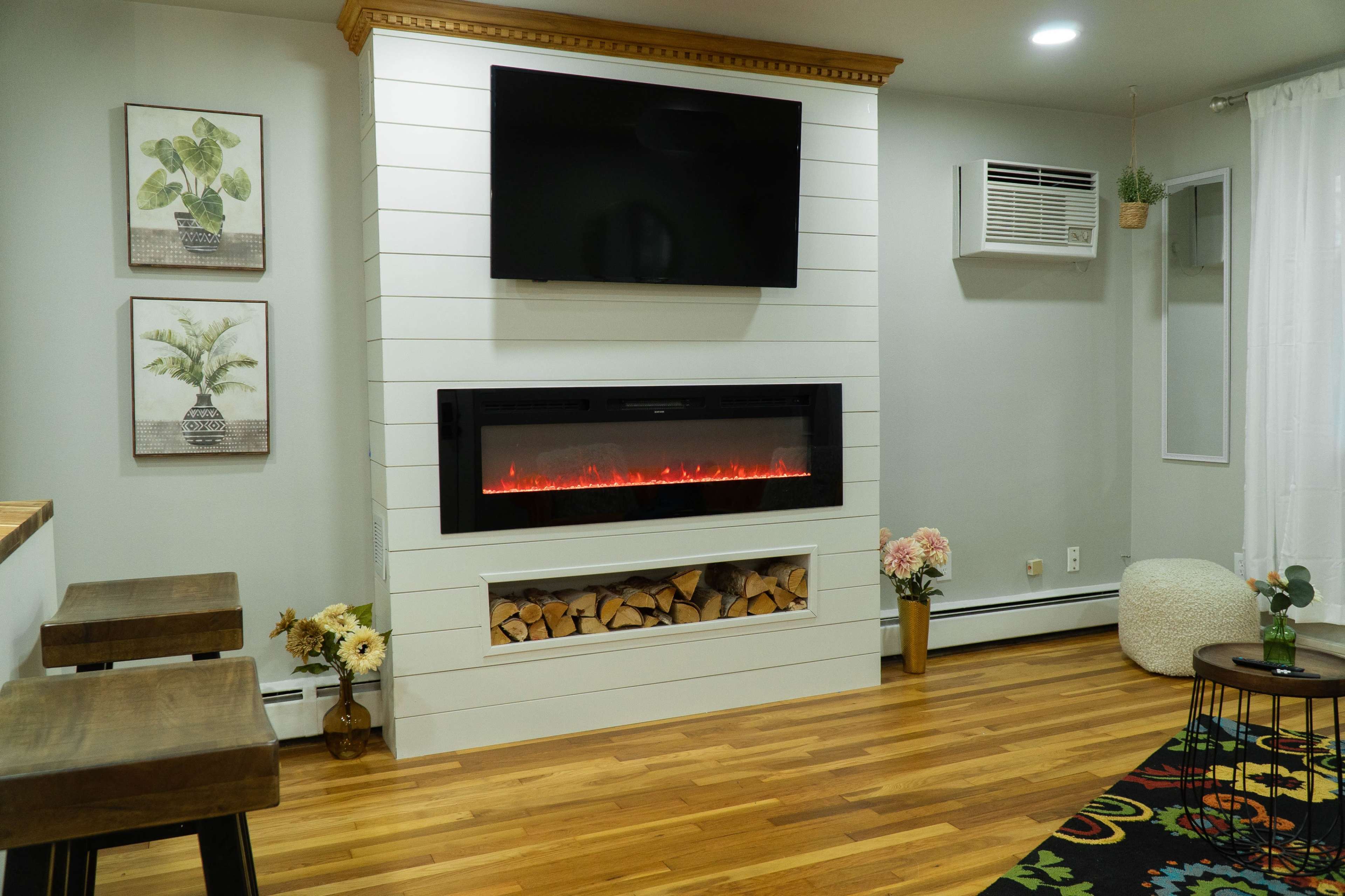 A modern living room with a wall-mounted television above an electric fireplace, surrounded by potted plants and decorative artwork.