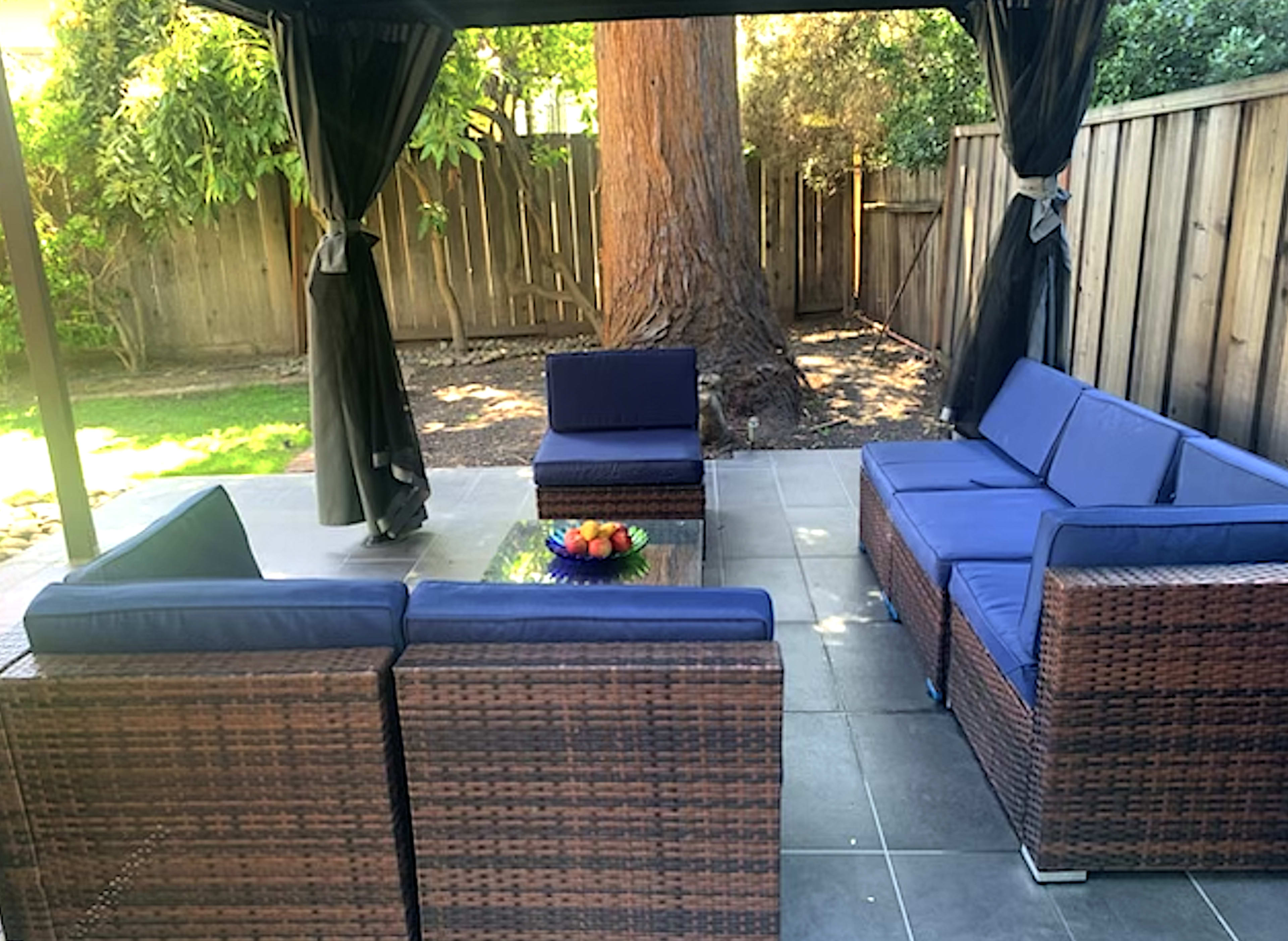 A patio area features a seating arrangement of wicker furniture with blue cushions under a canopy, alongside a fruit bowl on a glass table.