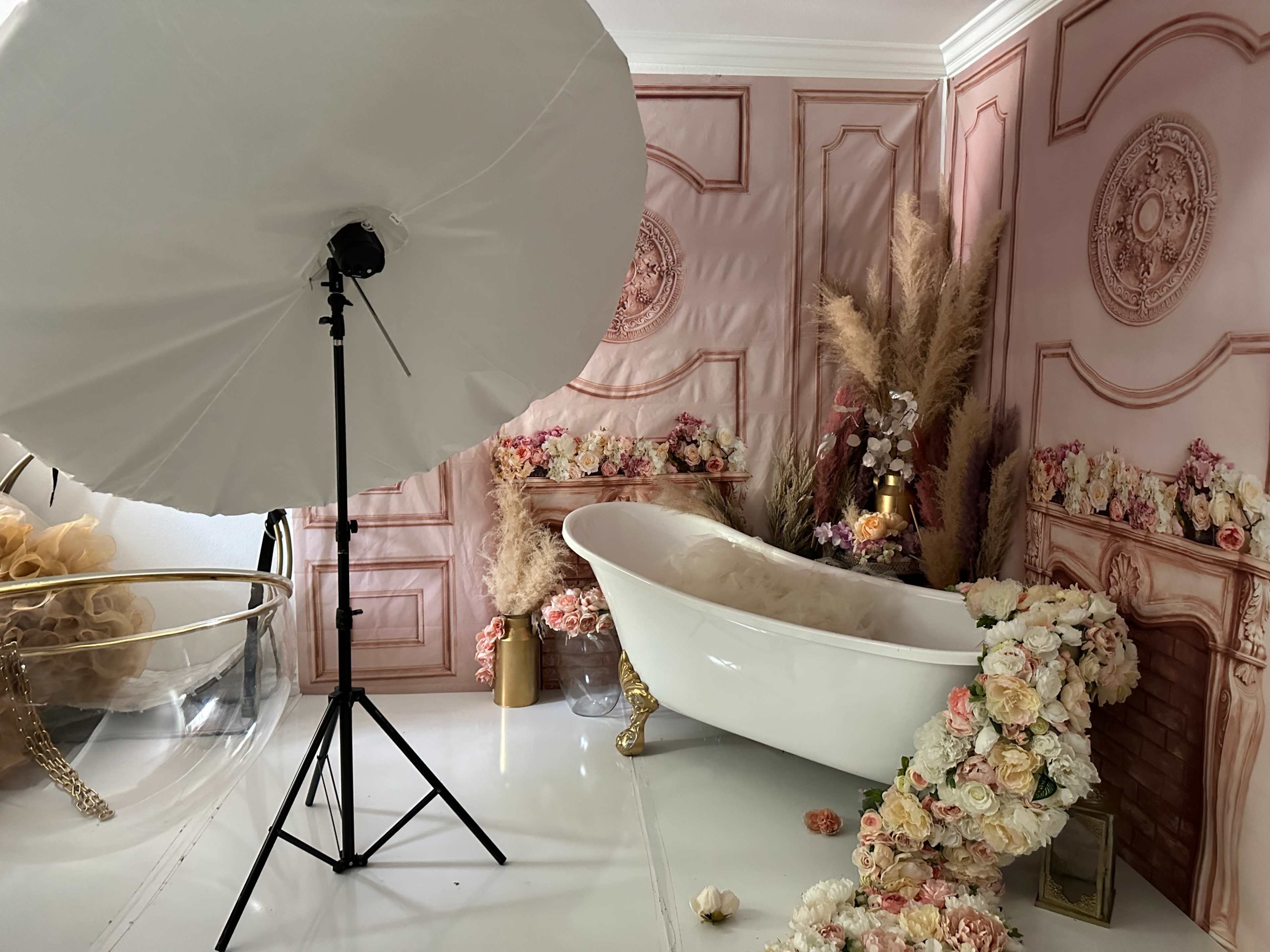 The image shows a decorated bathroom featuring a white clawfoot bathtub surrounded by floral arrangements and a large photography umbrella.