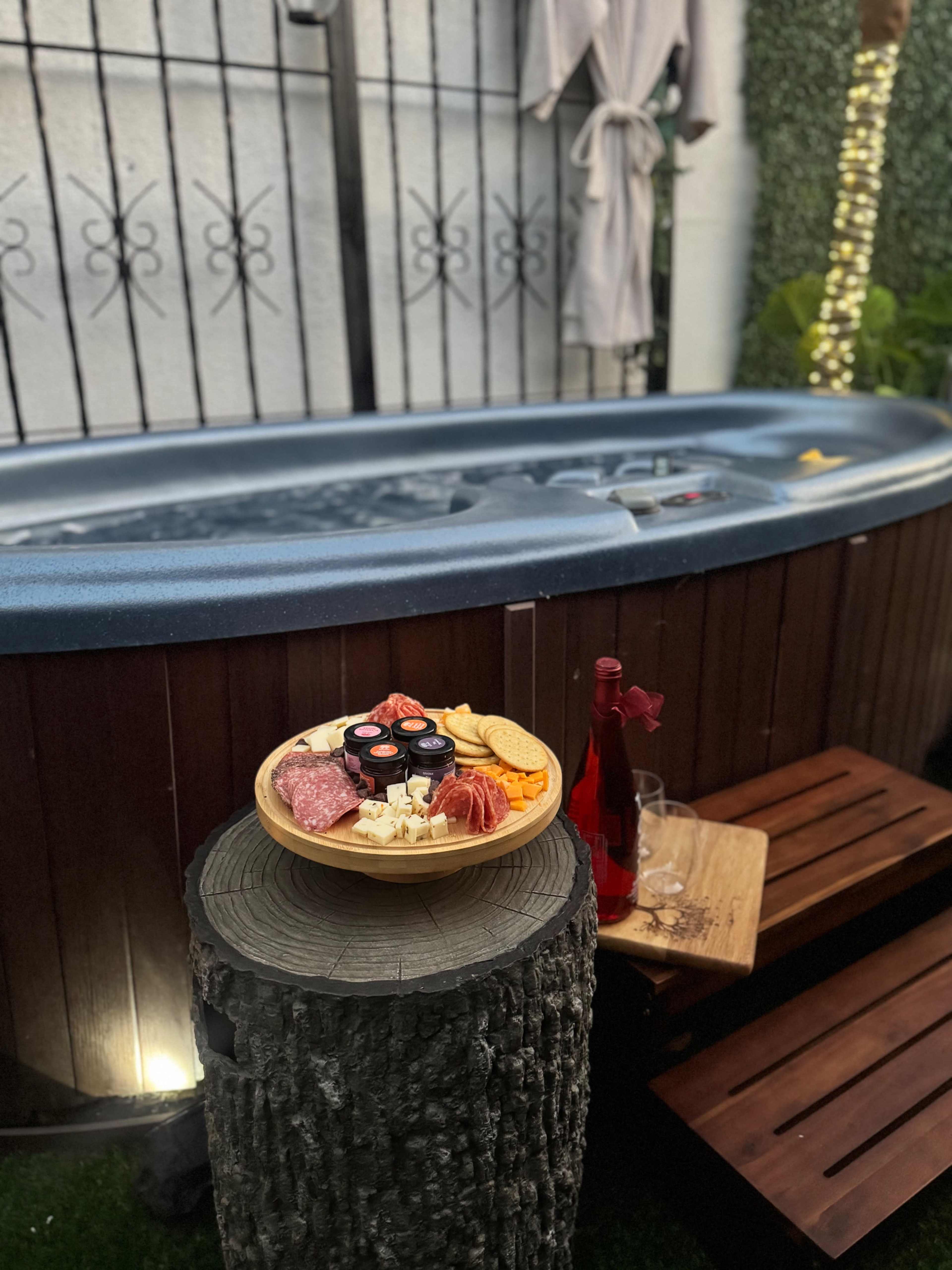 A hot tub is surrounded by greenery, with a wooden stool displaying a platter of various cured meats and cheeses, alongside a bottle of drink and glass.