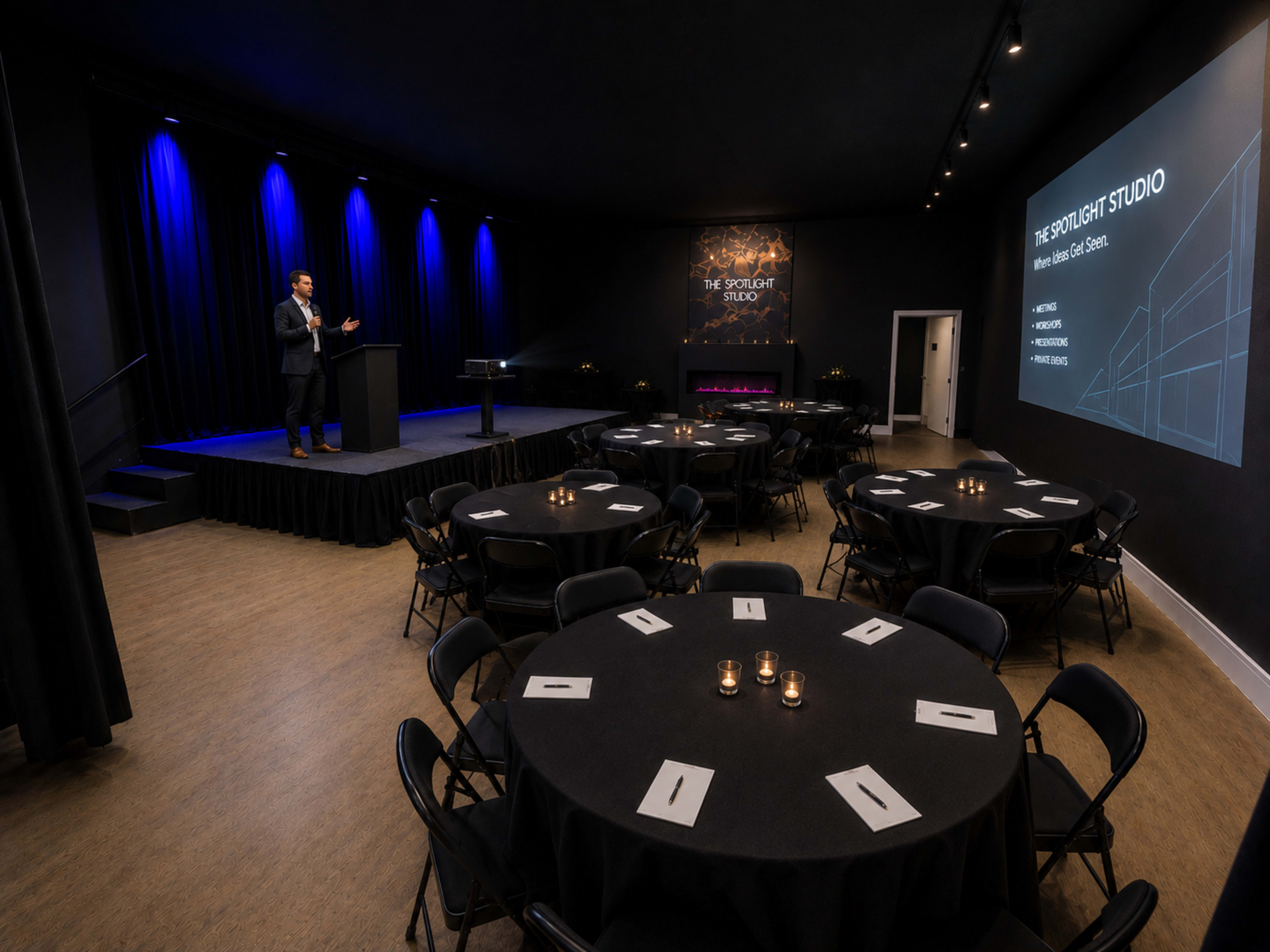 A speaker stands at a podium in a dimly lit event space with round tables set for guests and blue lighting accentuating the stage area.