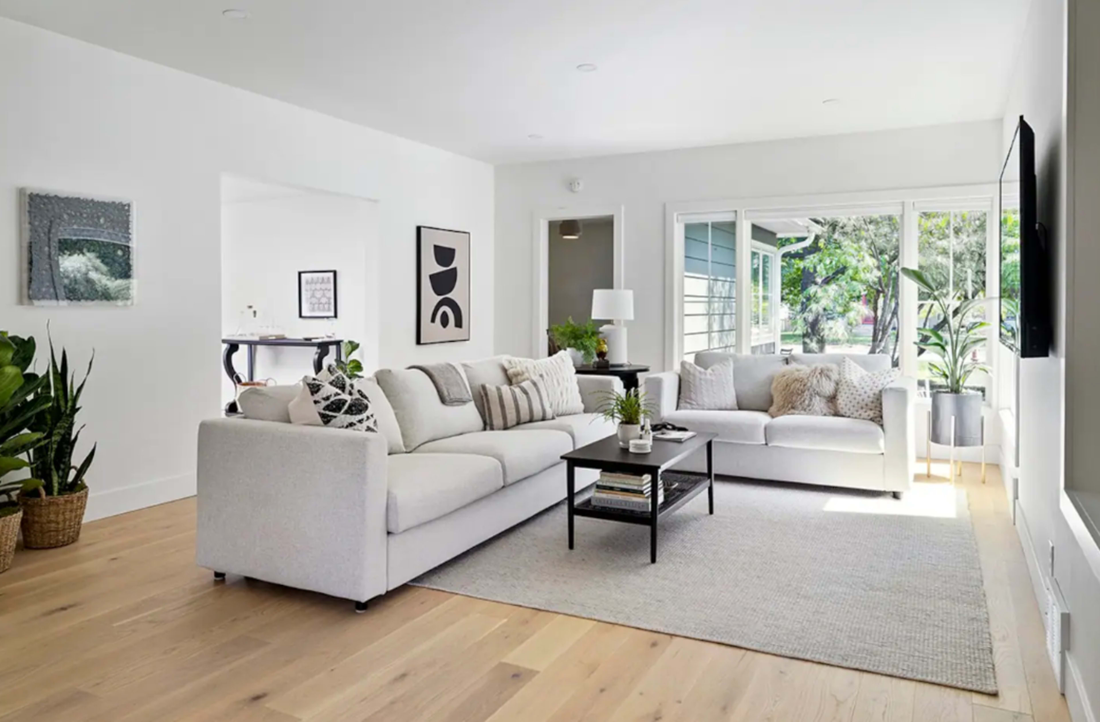 A bright living room features a large white sectional sofa, a black coffee table, and plants beside large windows that allow natural light to fill the space.