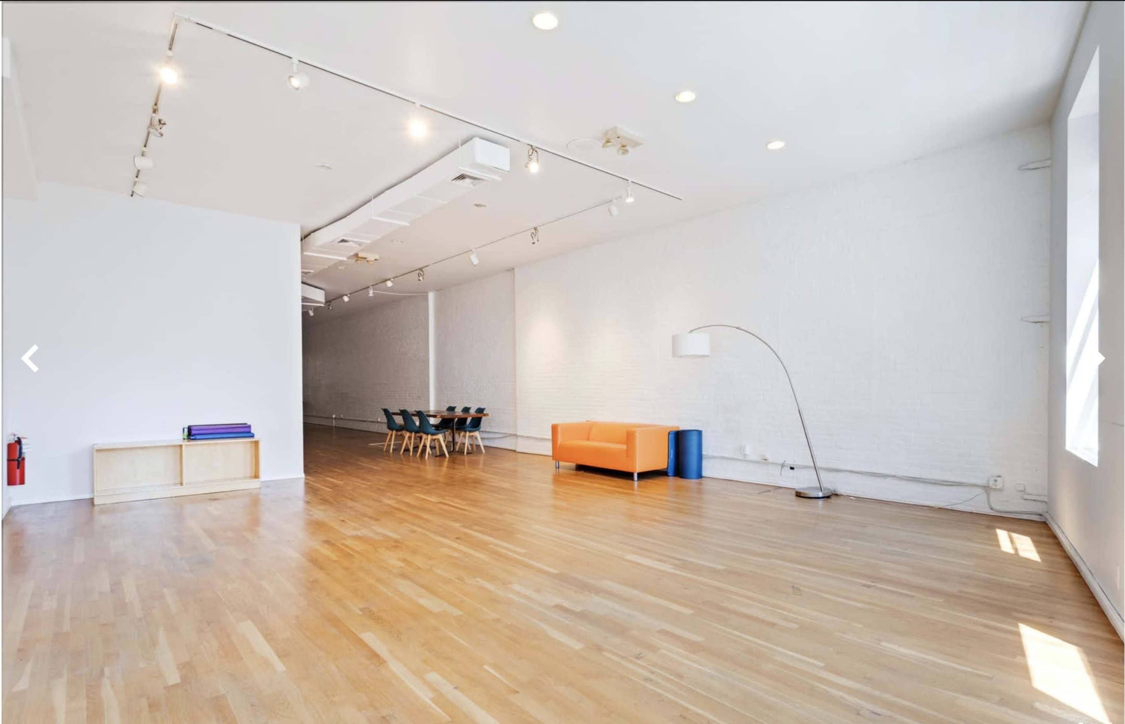 A spacious room with wooden flooring, white walls, a seating area featuring an orange couch, and a table with chairs in the background.