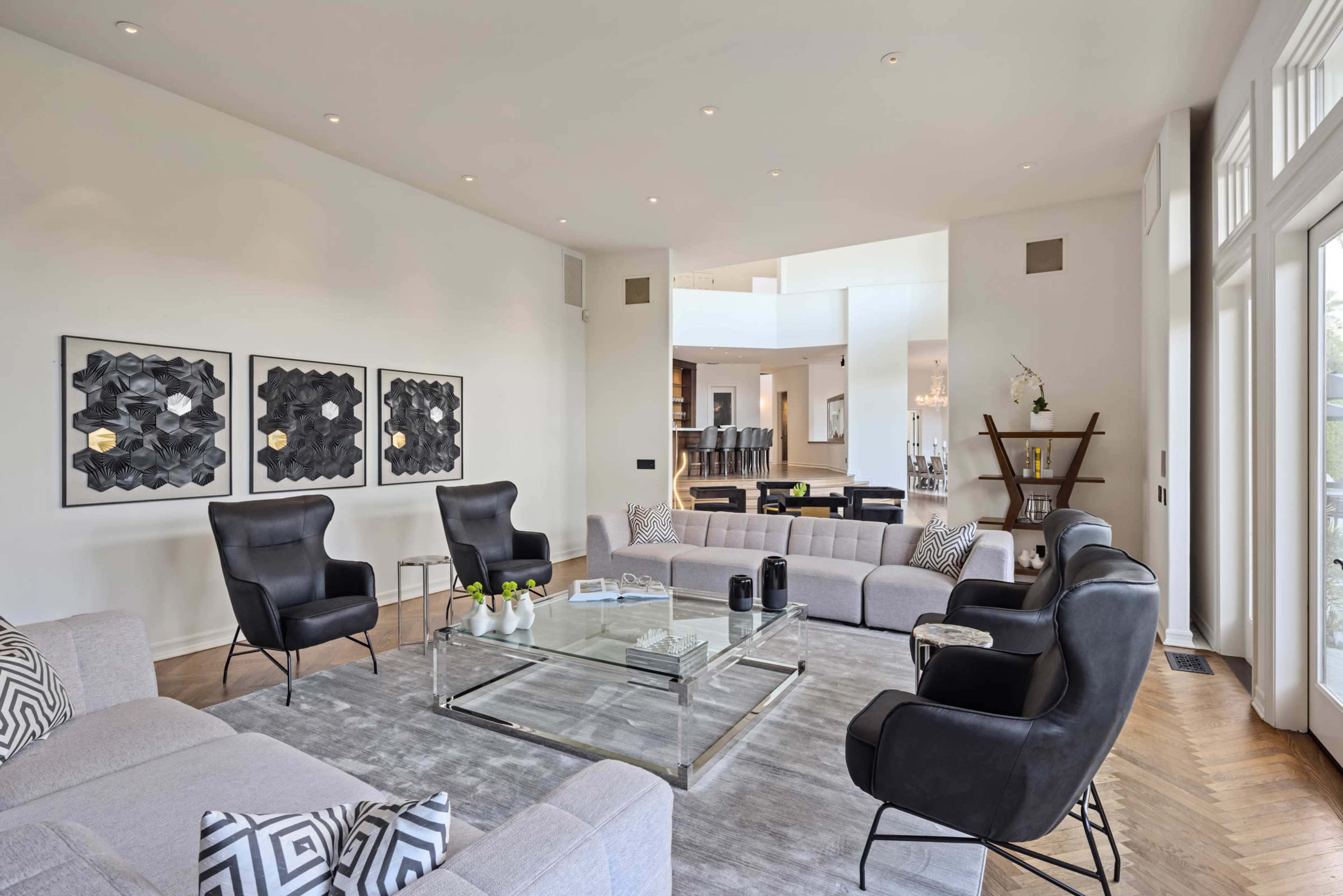 A modern living room features two black lounge chairs, a gray sectional sofa, a glass coffee table, and three framed artworks on the wall.