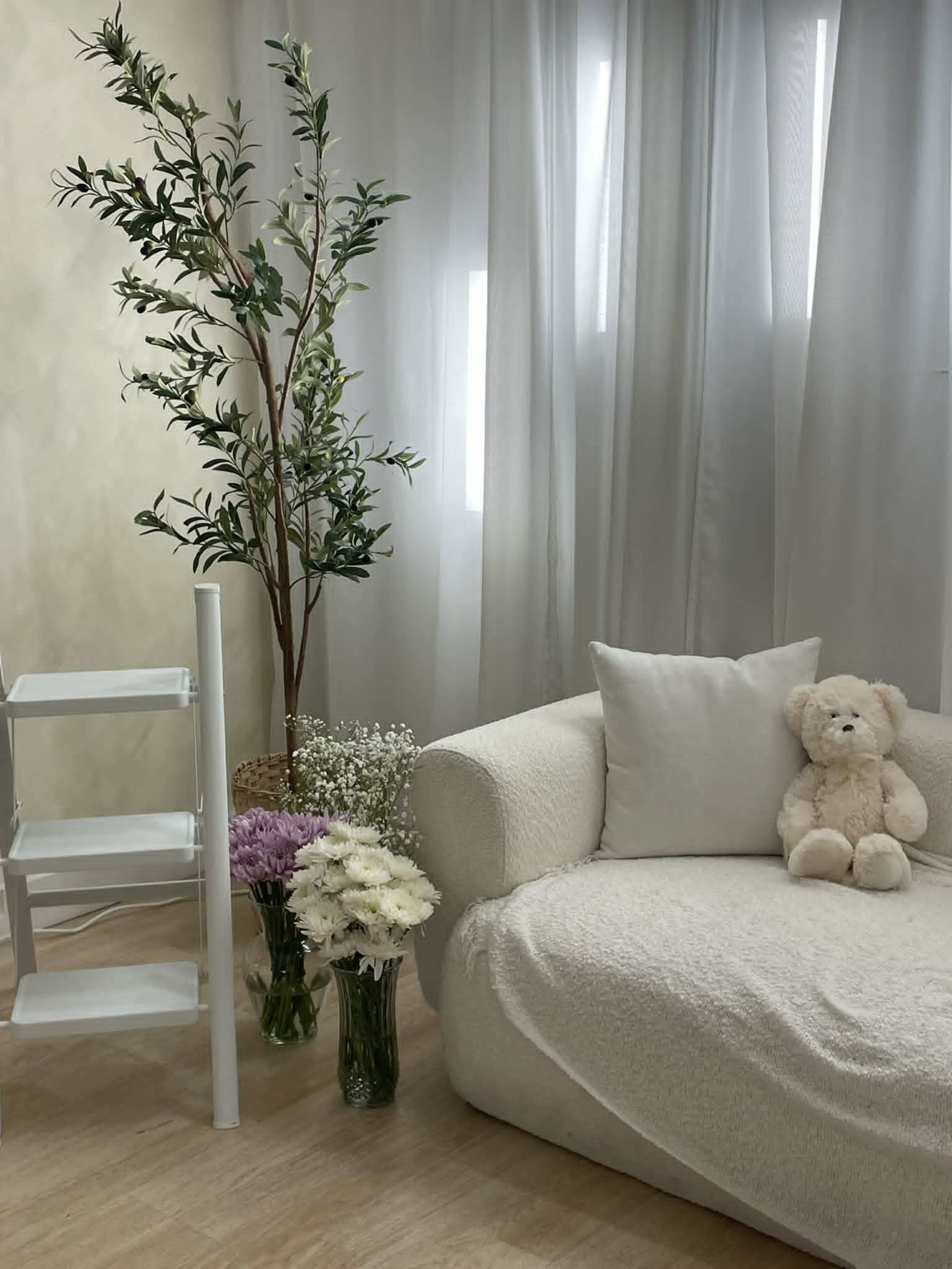 The scene features a cozy corner with a white couch draped in a blanket, a soft teddy bear, a shelf, and several flower arrangements beside a potted plant.