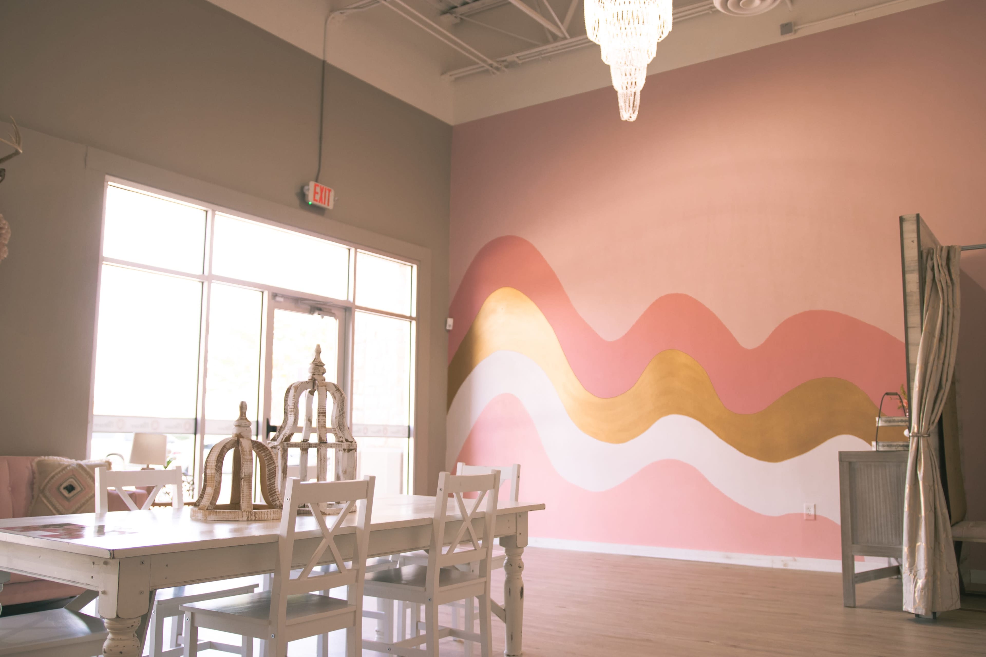 A bright interior features a dining table with white chairs and a colorful wave mural on the pink wall.